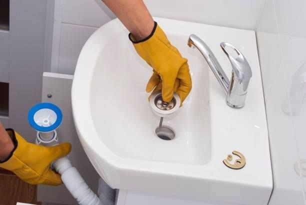 Plumber in yellow gloves repairs a white sink, holding the drain stopper. Close up shows plumbing.
