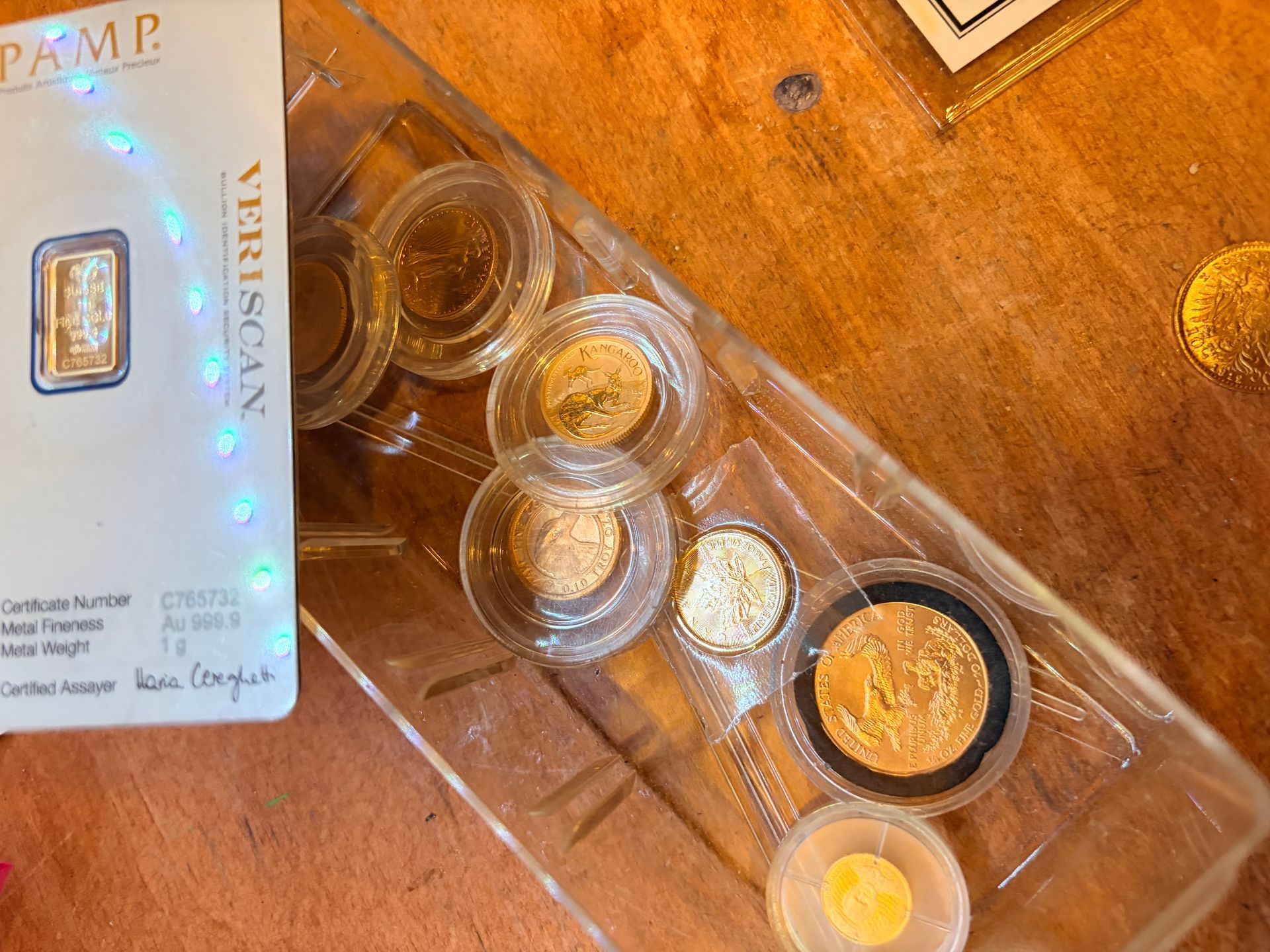 A 1-gram PAMP gold bar in a plastic package next to a tray containing six small gold coins on a wooden surface.