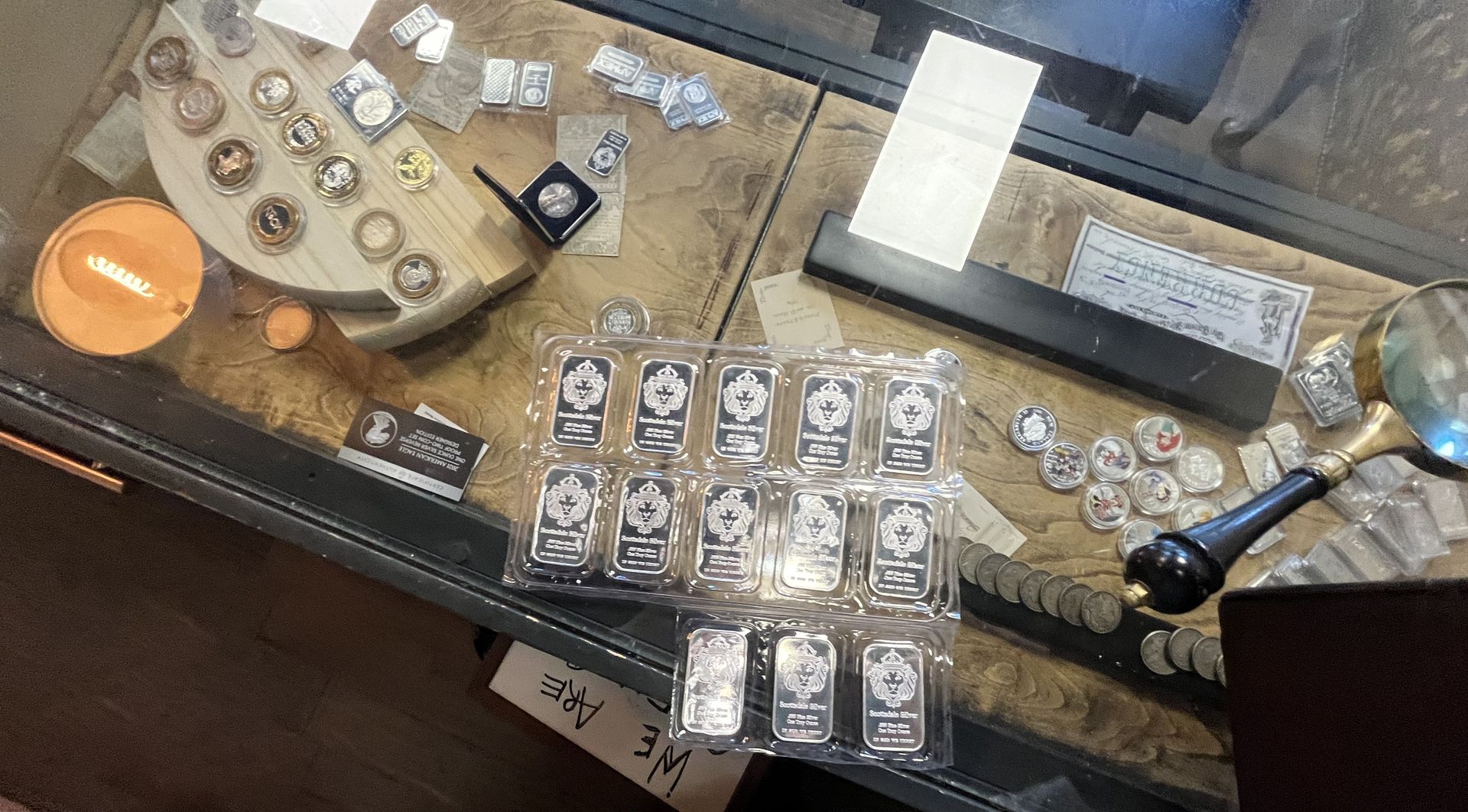 A collection of silver coins and rectangular bullion bars displayed on a wooden surface with a magnifying glass.