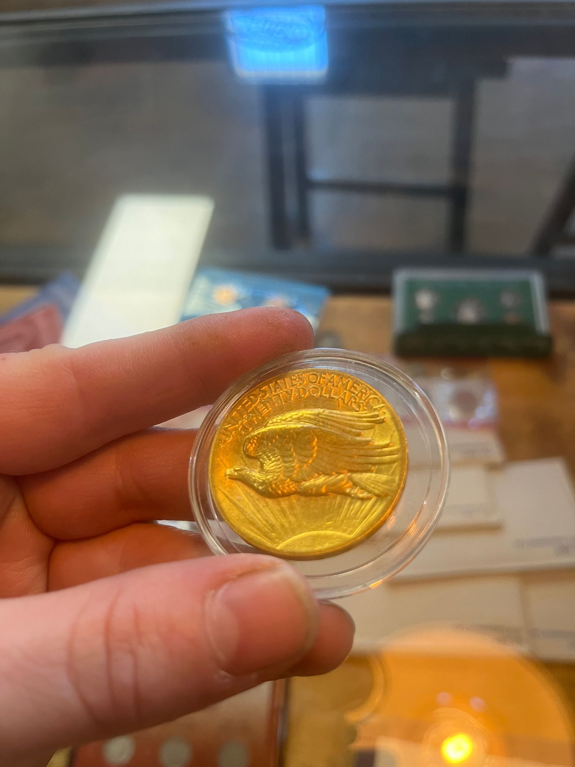 A hand holds a gold coin in a clear plastic protective case, showing an eagle design against a blurred indoor background.