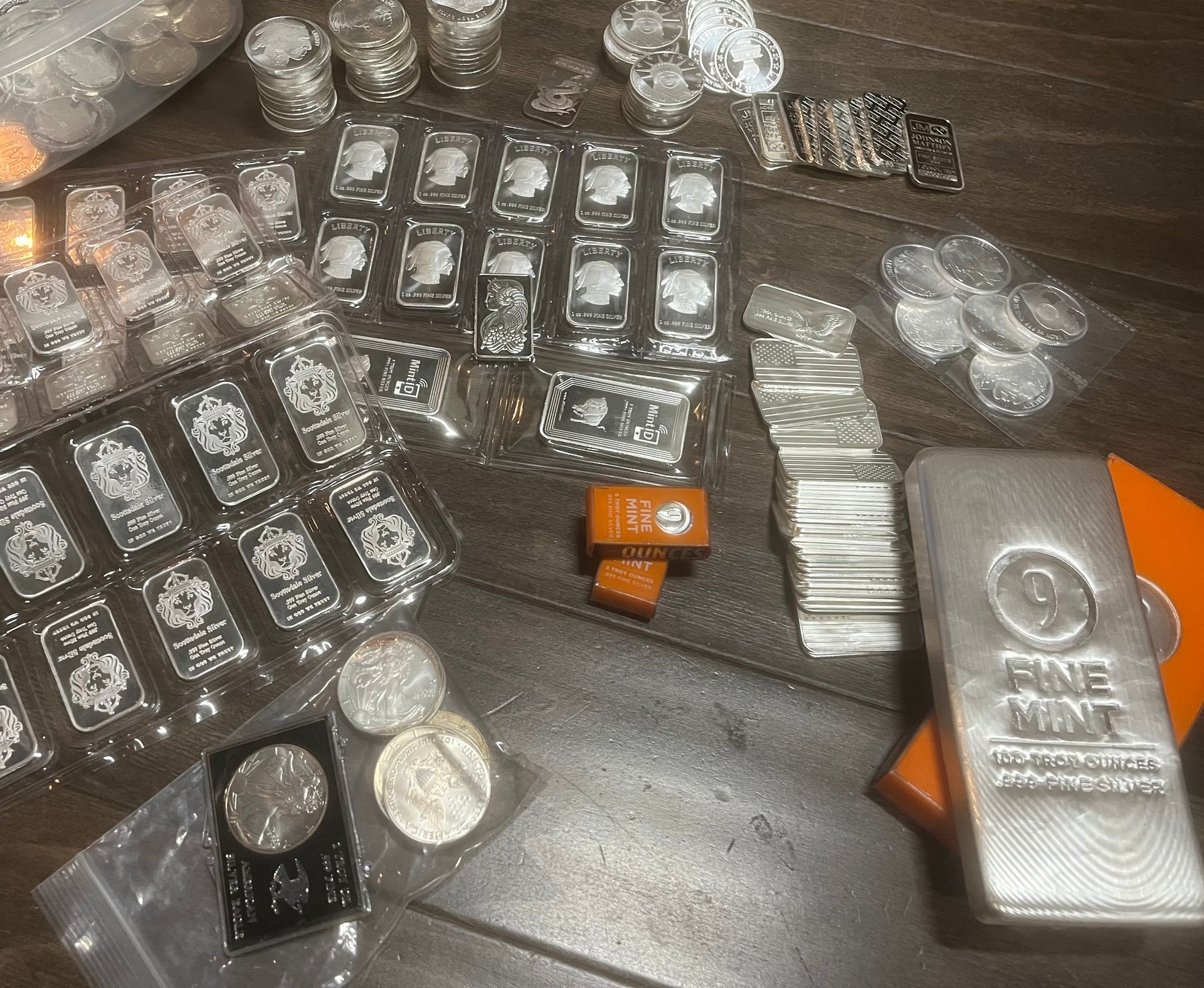 A large collection of silver bars and coins of various sizes arranged on a dark wooden surface.