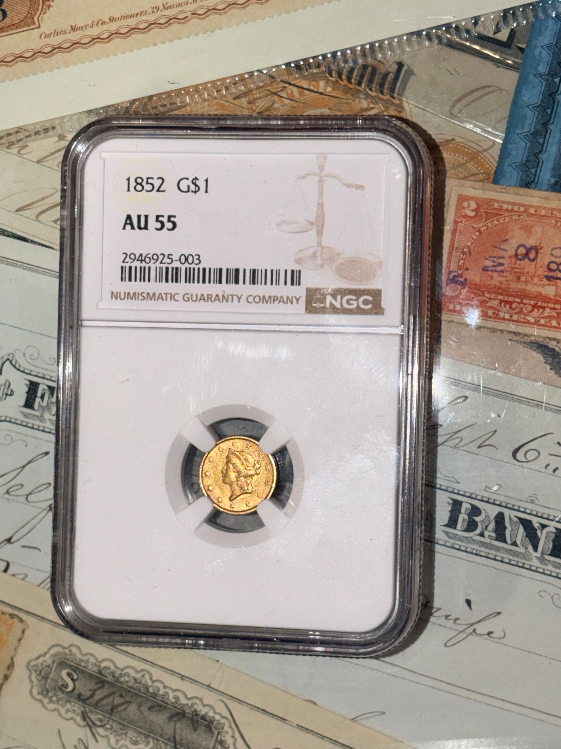 An 1852 gold dollar coin in an NGC protective slab graded AU 55, resting on a collection of vintage paper currency.
