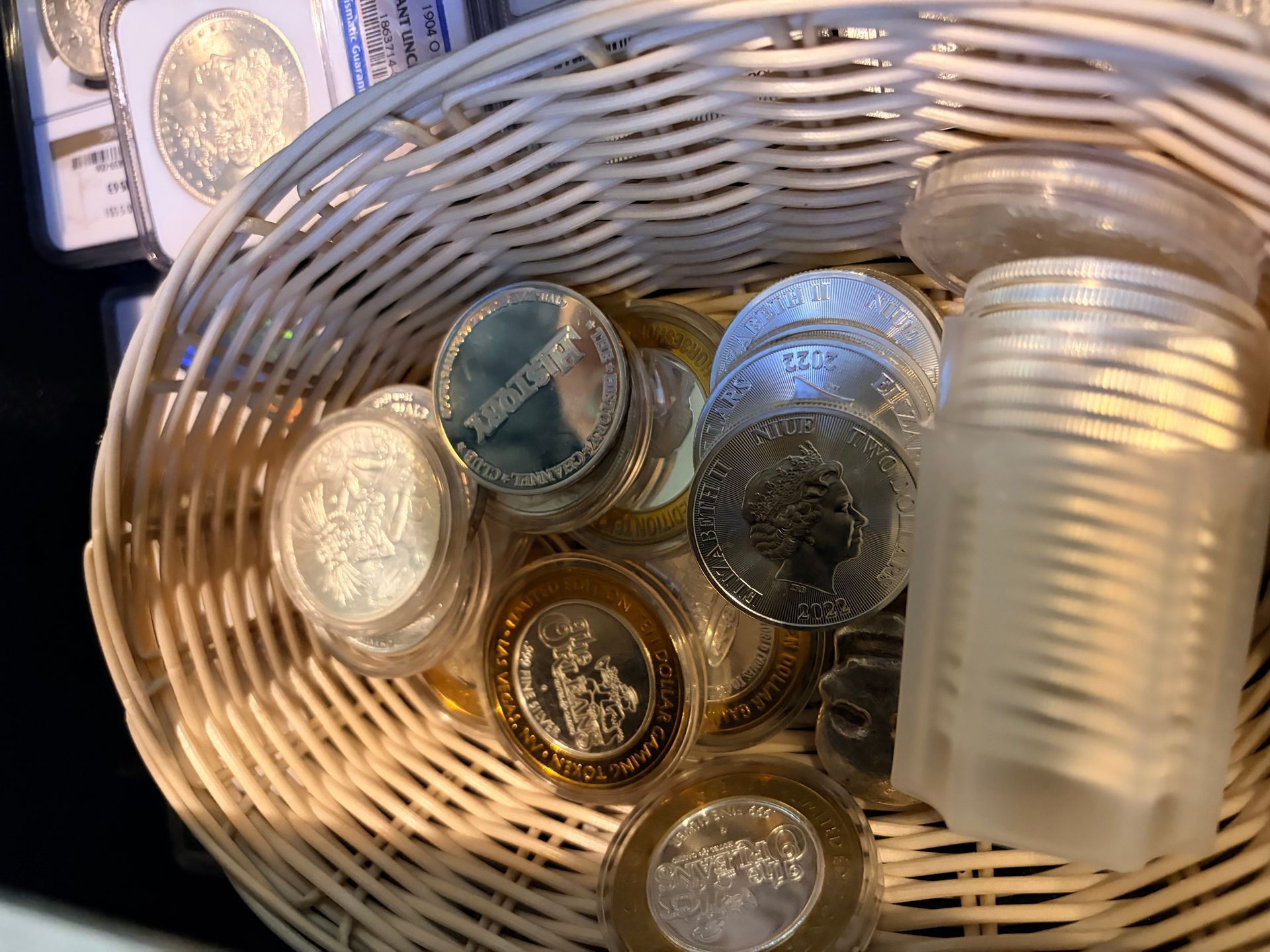 A wicker basket filled with various silver coins, including several in protective plastic capsules and a stack in a tube.