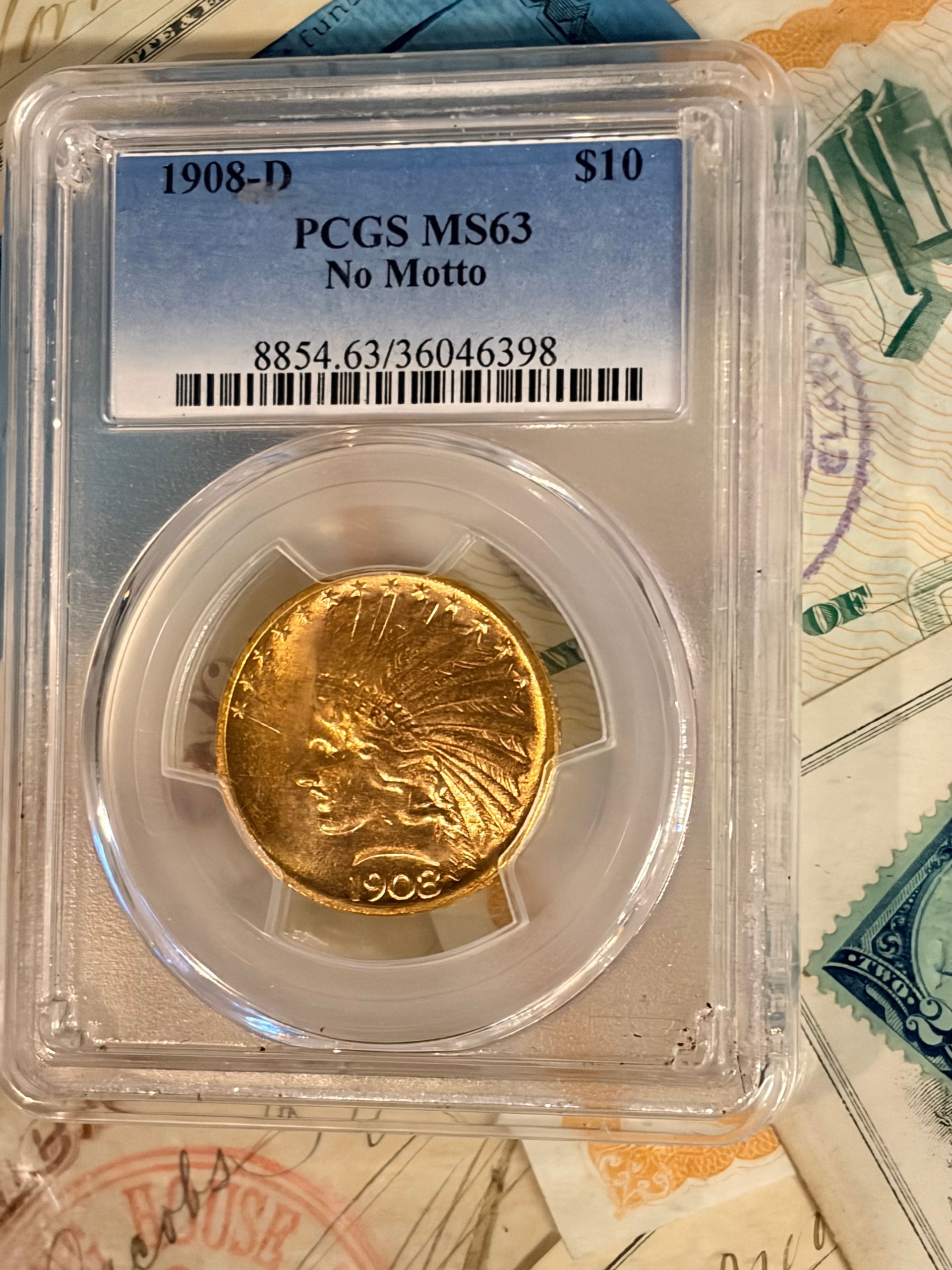 A 1908-D $10 Indian Head gold coin graded MS63 by PCGS, housed in a plastic slab, resting on paper currency.