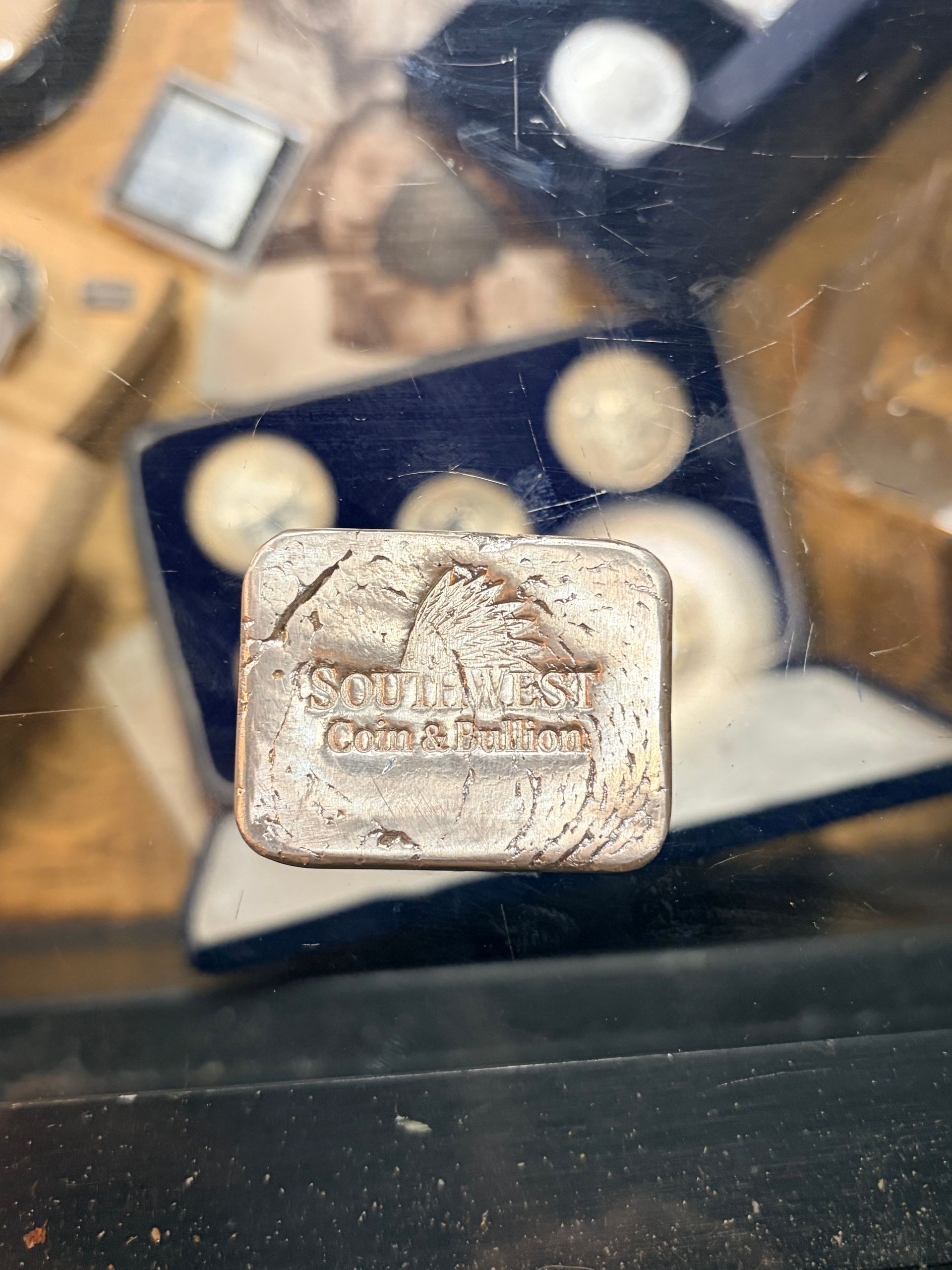 A rectangular, silver-colored metal bar with stamped text, resting on a surface near a blue-lined coin display case.