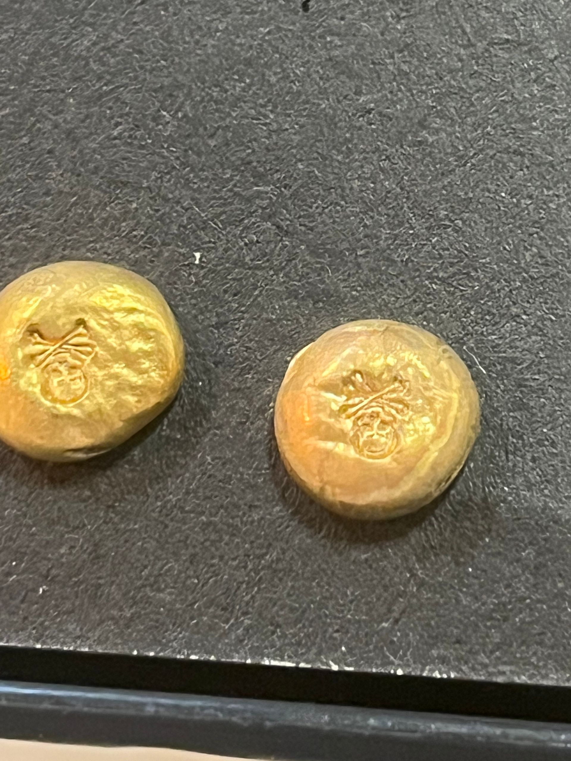 Two small, circular gold-colored coins or tokens with an embossed emblem of a stylized butterfly or bug on a dark surface.