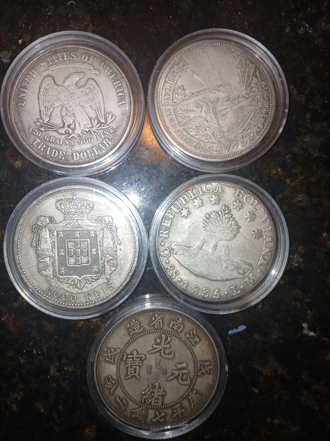 Five silver coins in protective circular capsules arranged on a dark, textured surface.