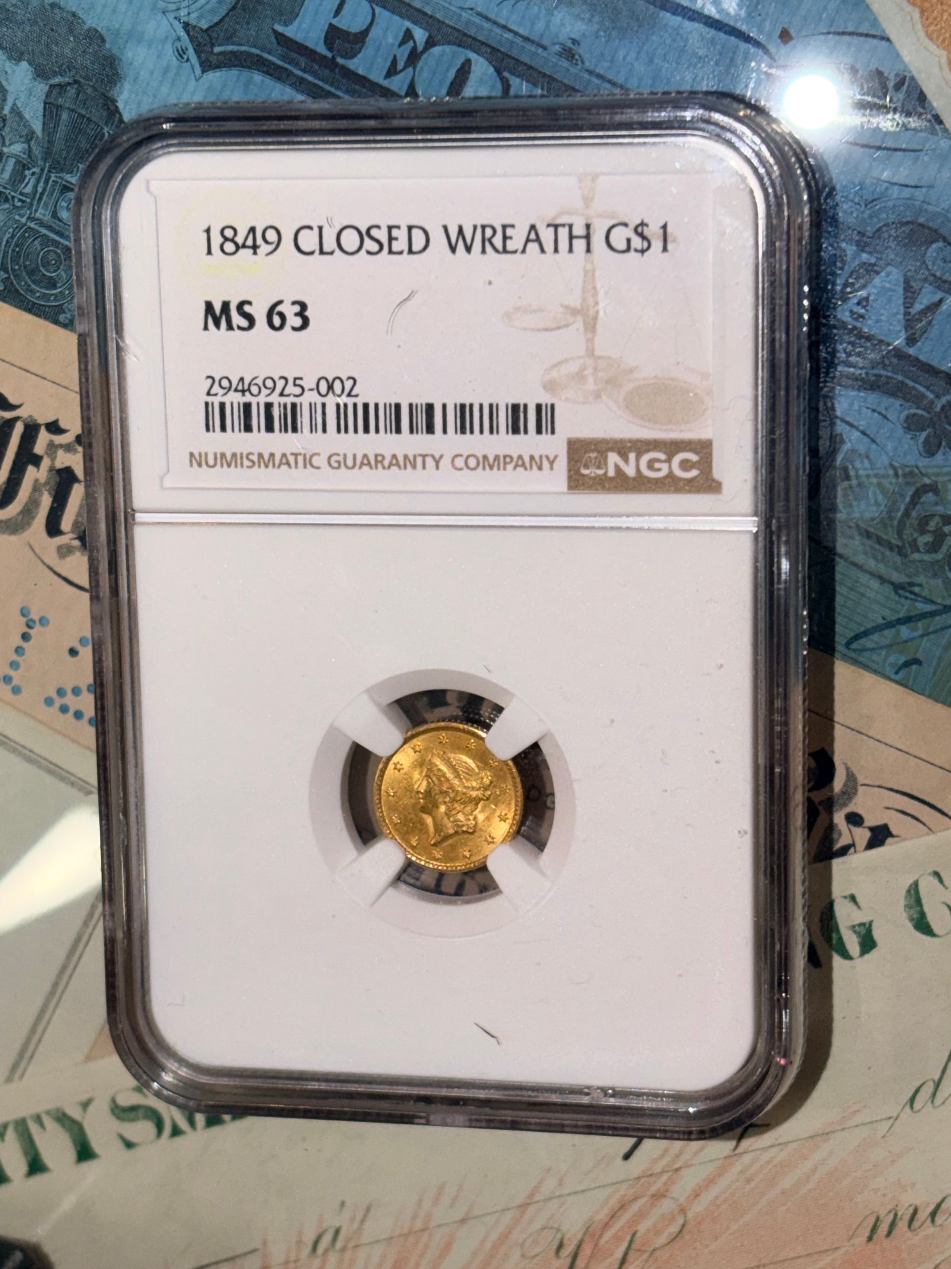 An NGC-certified 1849 US gold dollar coin in an MS 63 slab holder, resting on a background of antique currency.