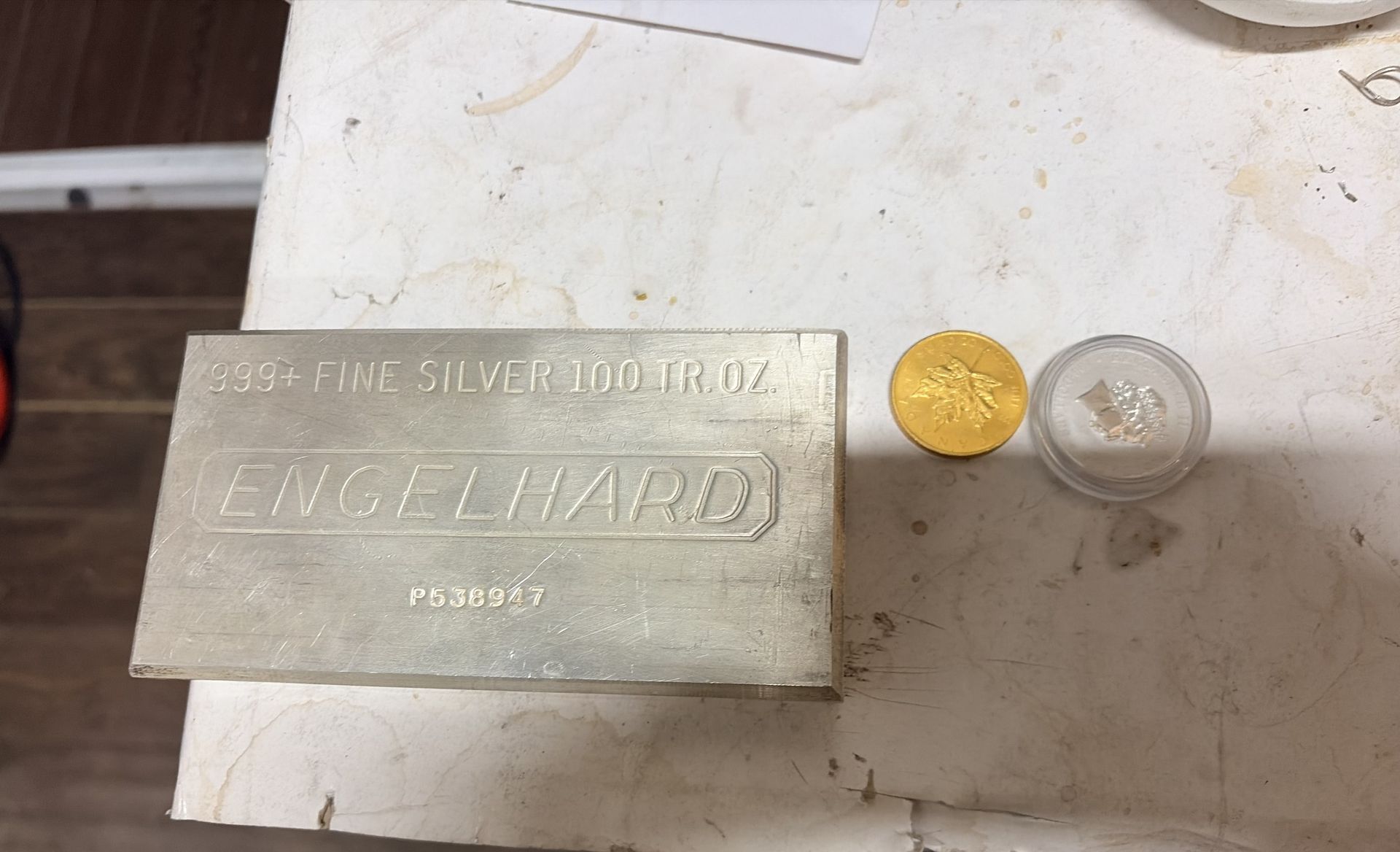 A 100 troy ounce silver Engelhard bar sitting next to a small gold coin and a small silver coin on a white surface.