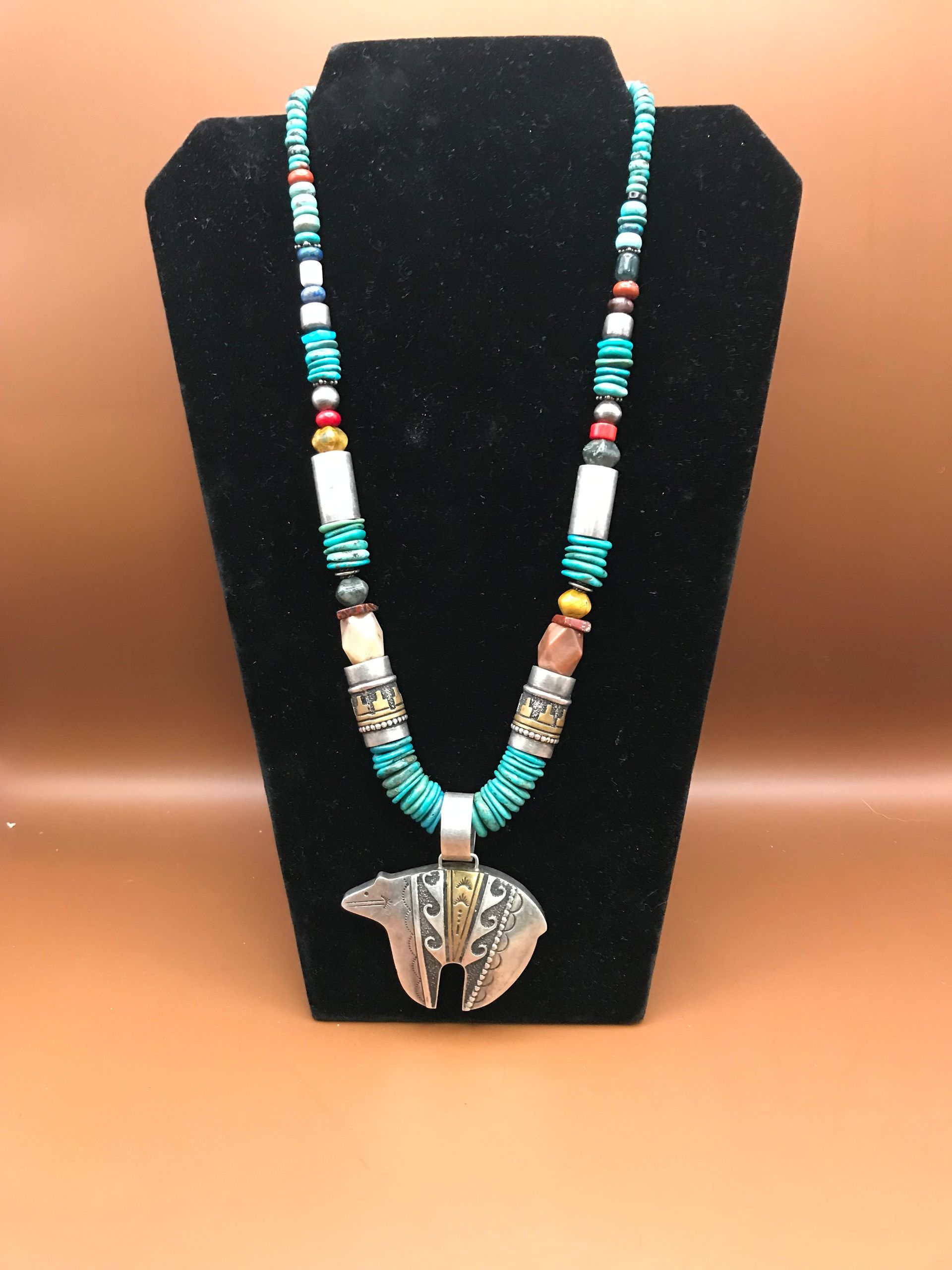 A turquoise, white, and red beaded necklace with a silver bear-shaped pendant centered on a black display stand.