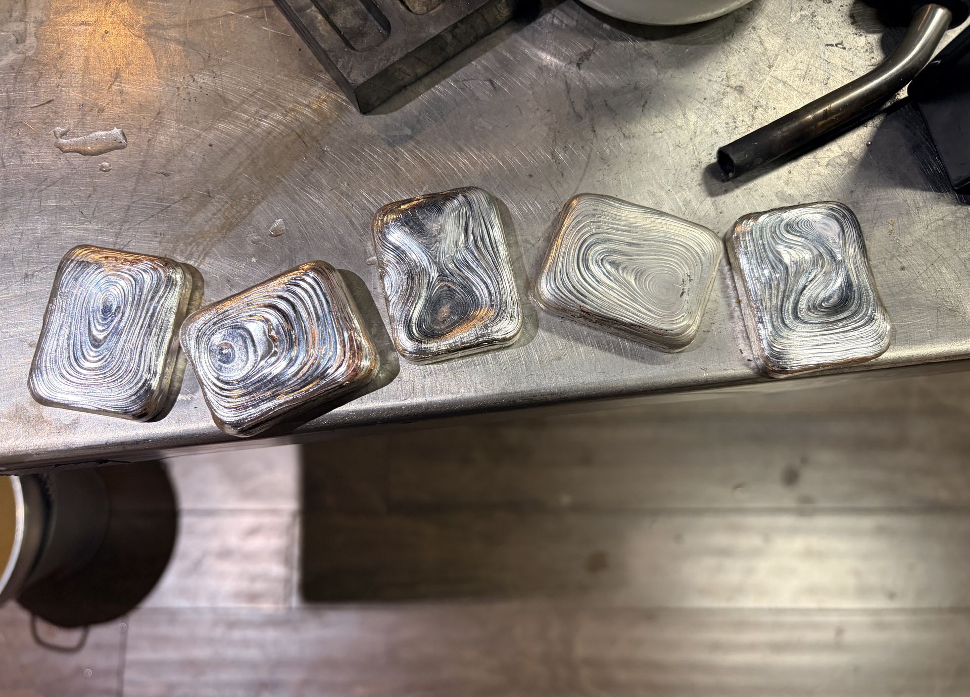 Five rectangular silver bars with swirling, cooling patterns sitting on a metal workbench.