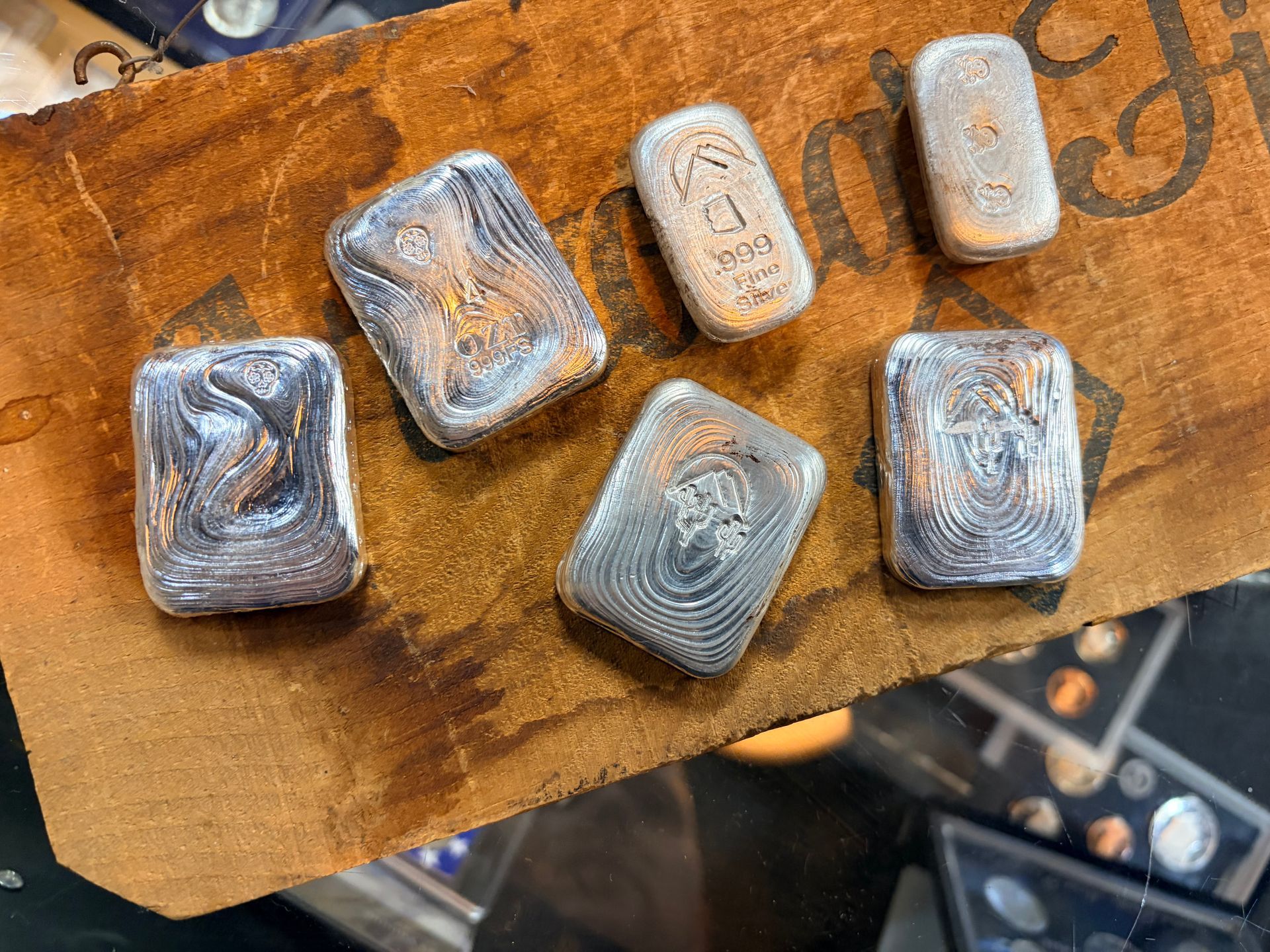 Six small silver bars with embossed designs arranged on a textured wooden surface.