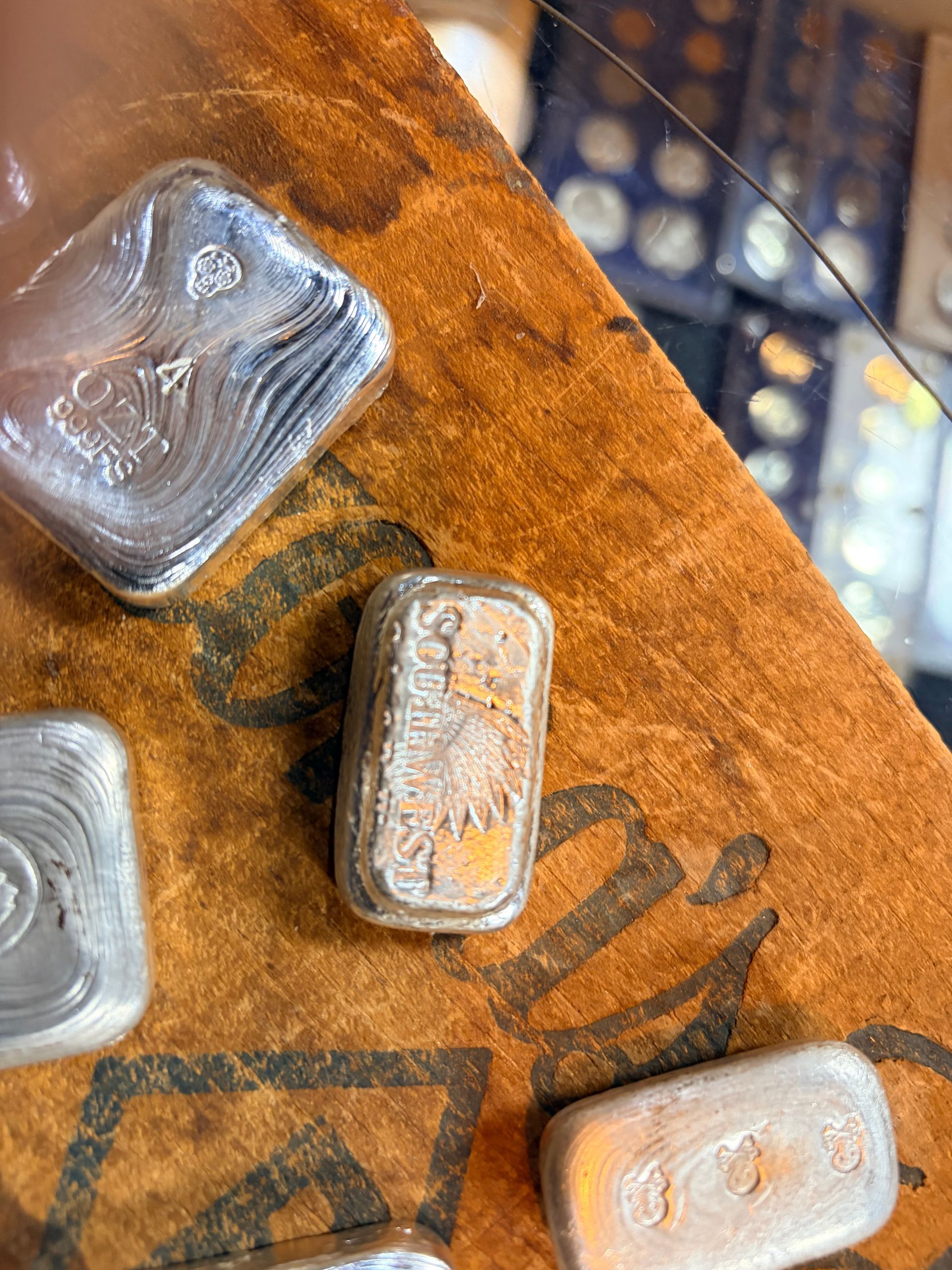 Several small rectangular silver bars resting on a wooden surface with blurred coin collection holders in the background.