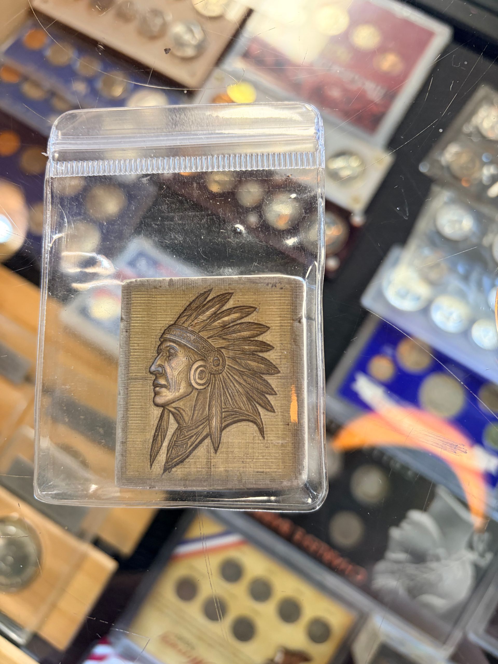 A square metal ingot featuring an embossed Native American head in profile, sealed in a clear plastic bag over coin sets.