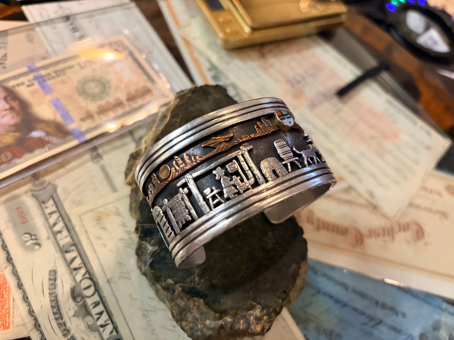 A silver cuff bracelet with intricate desert scene cutouts rests on a dark, rough rock amidst papers and a gold scale.
