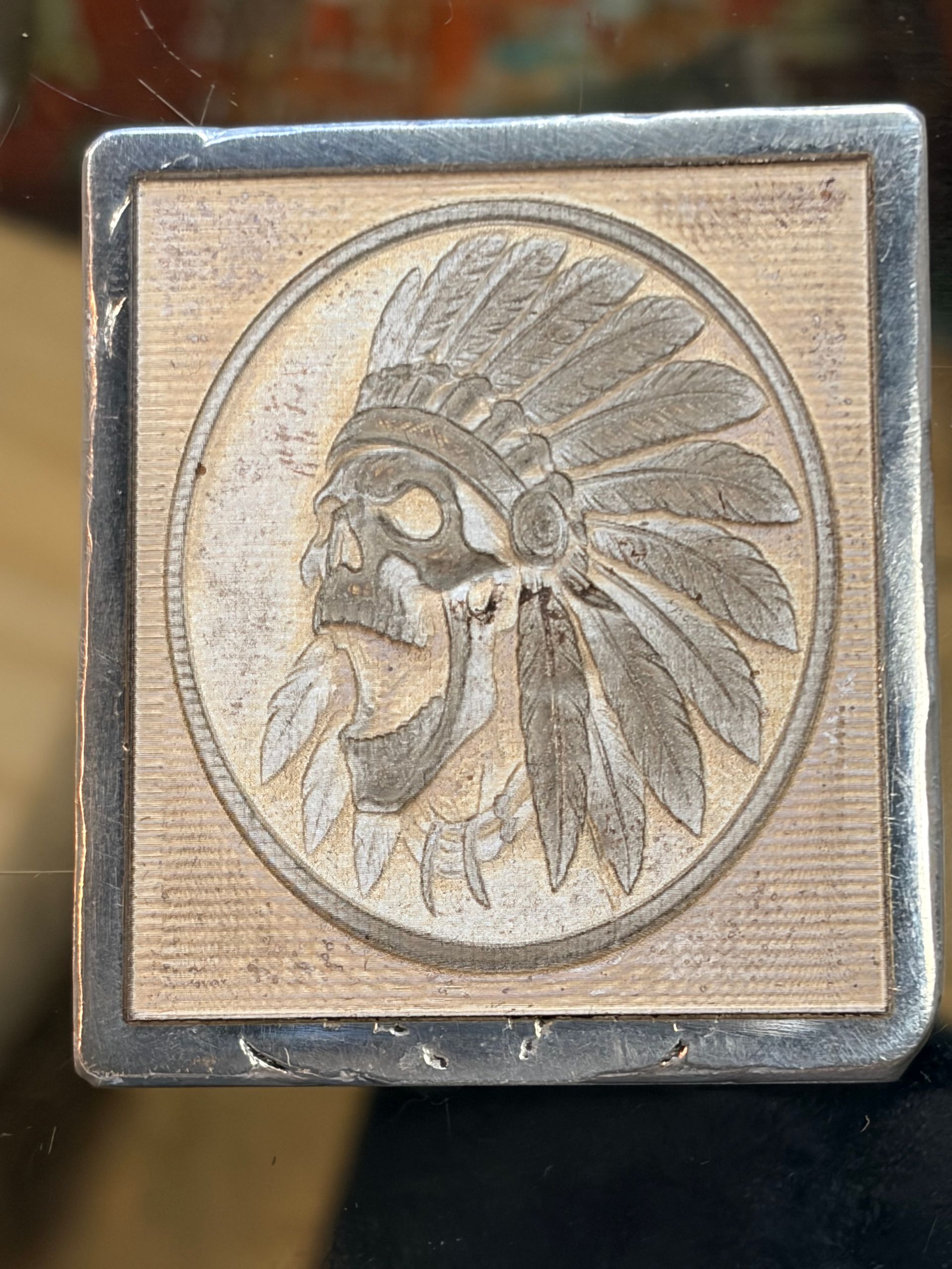 A square, metallic relief carving of a skull wearing a feathered headdress, centered within a circular border.