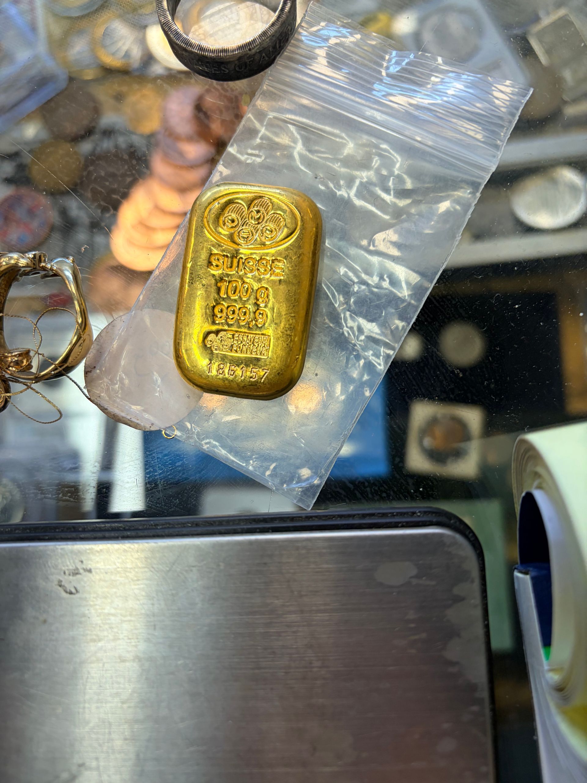 A small, gold rectangular ingot in a clear plastic bag, resting on a silver surface near other miscellaneous coins.