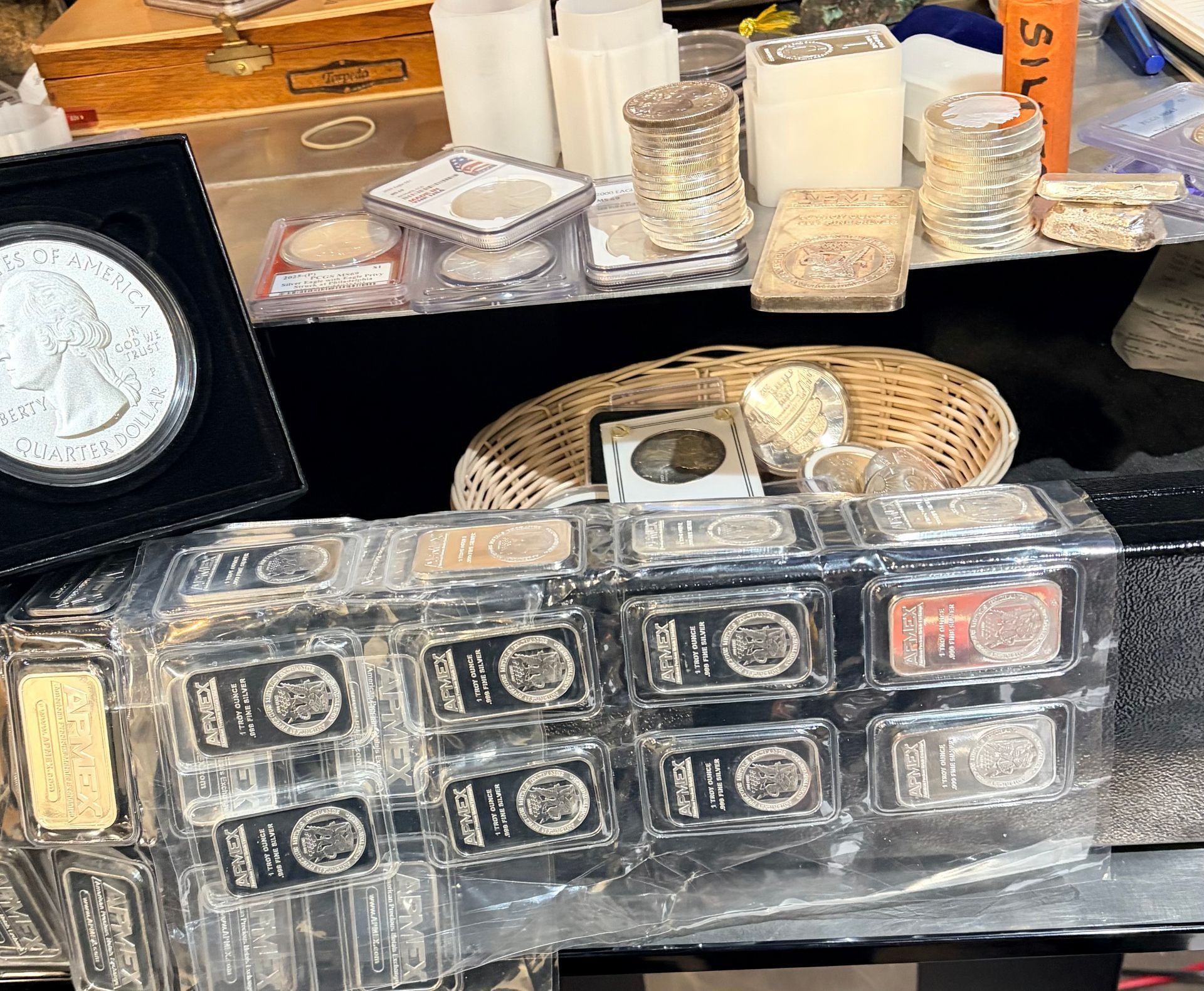 A collection of silver coins, bars, and bullion stored in plastic cases and a basket on a table.