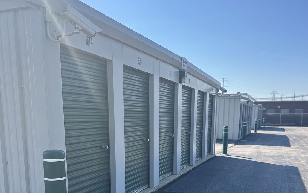 A row of white self-storage units with green roll-up doors under a clear blue sky.