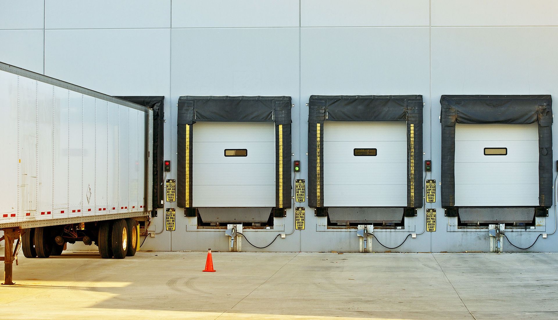 A white semi-truck parked at a warehouse loading dock with three empty loading bays and a single orange traffic cone.
