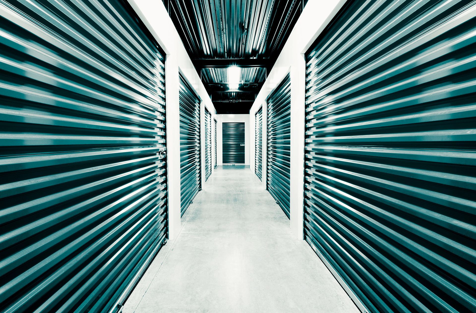 A long, narrow hallway in a self-storage facility featuring rows of green, corrugated metal rolling doors and a concrete floor.