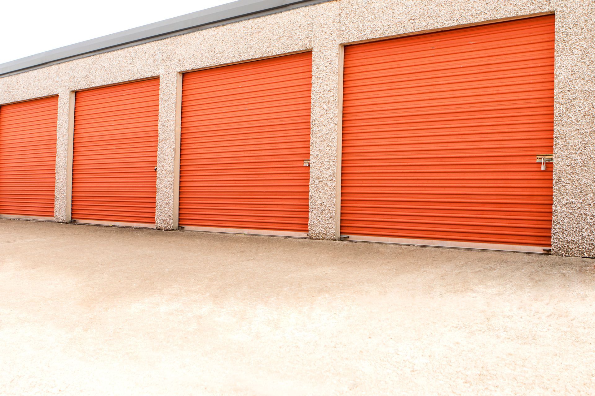 A row of four closed orange roll-up storage unit doors set into a textured beige building exterior.