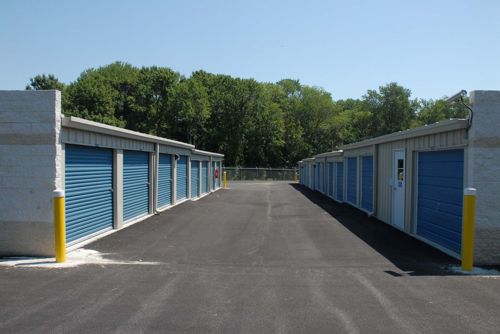 A paved driveway between two rows of storage units with blue roll-up doors, flanked by yellow safety bollards.