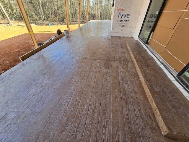 A freshly poured wood-plank stamped concrete patio deck under construction next to a house with Tyvek wrap.