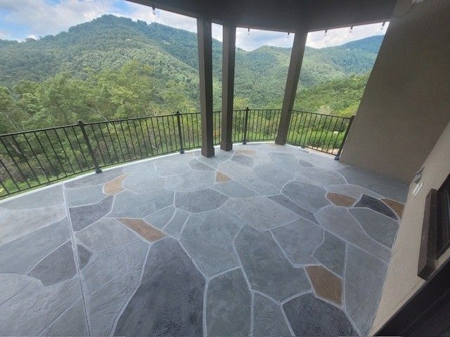 A balcony with a gray stone-patterned floor overlooking lush green mountains under a blue sky with white clouds.