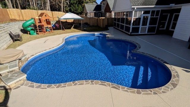An irregularly shaped backyard swimming pool with a stone border, adjacent to a patio, screen porch, and play area.