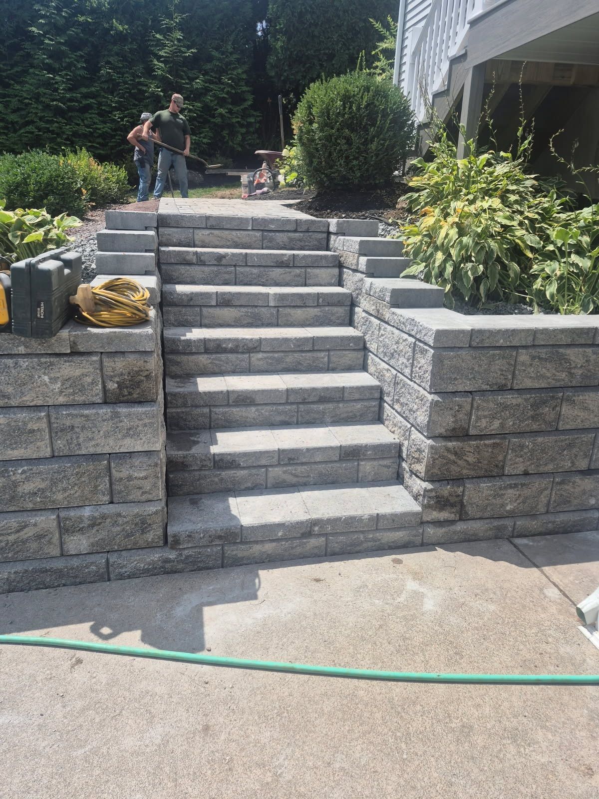 Stone steps and retaining walls leading to a patio, with two workers and garden plants in a backyard.