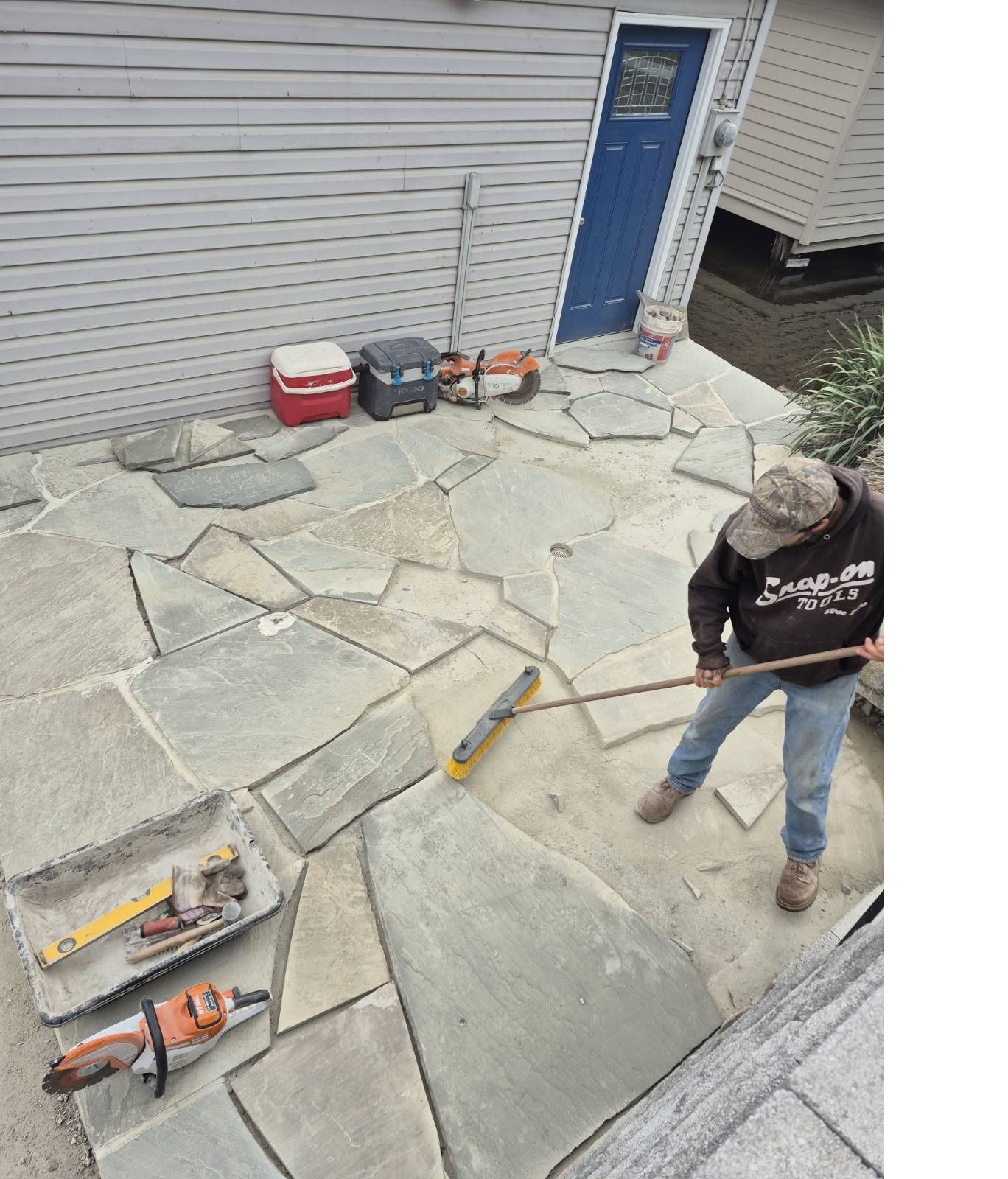 Worker installing stone pavers beside a house, with tools scattered on the patio.