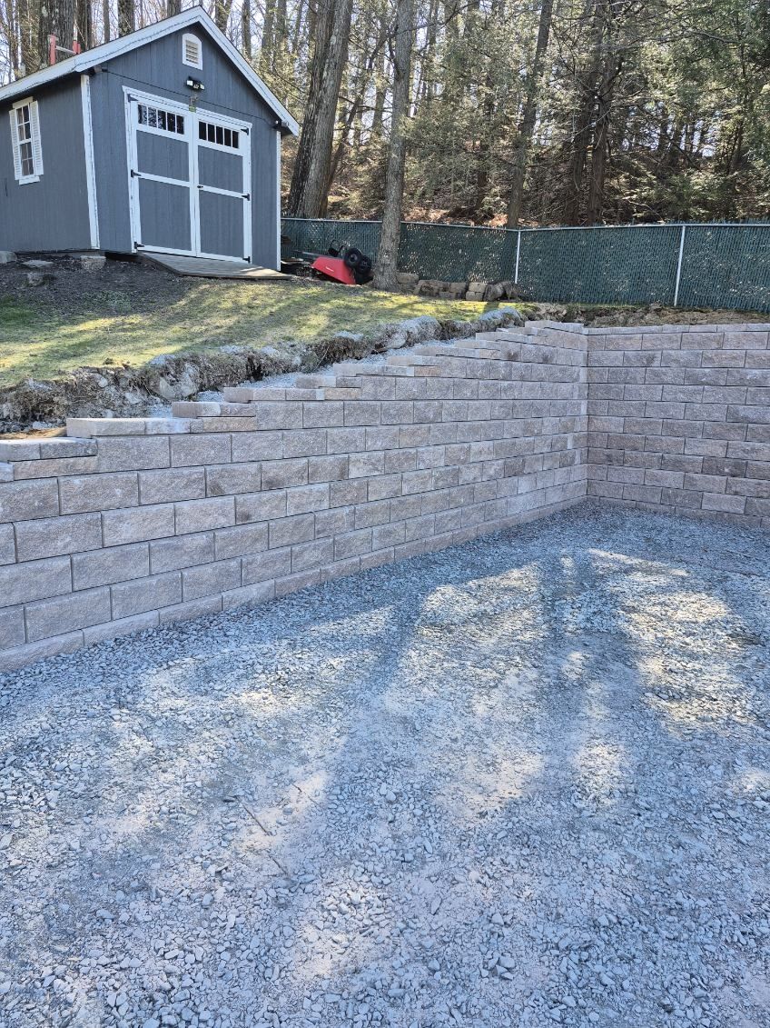 Stone retaining wall beside a gravel driveway, with a gray shed and wooded yard in the background.