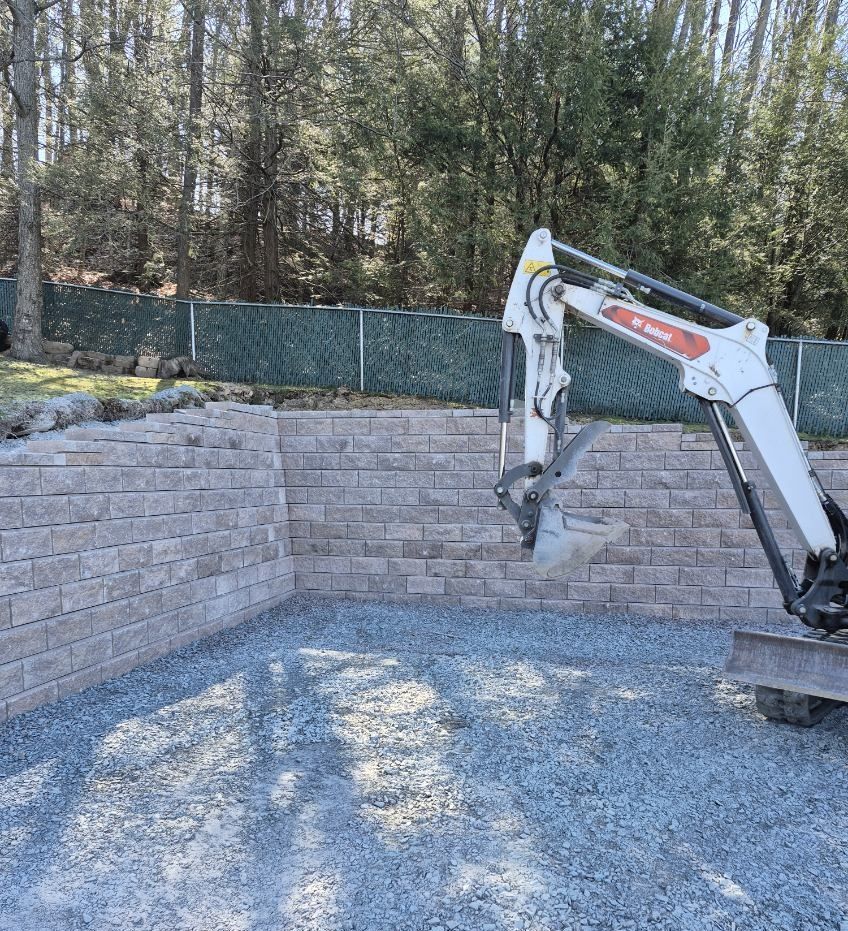 Excavator arm working over a gravel area beside a terraced stone retaining wall in a wooded yard