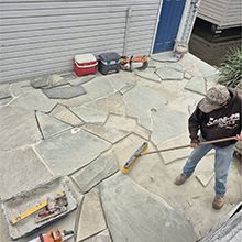 Worker laying irregular stone pavers in an outdoor patio area near a house.