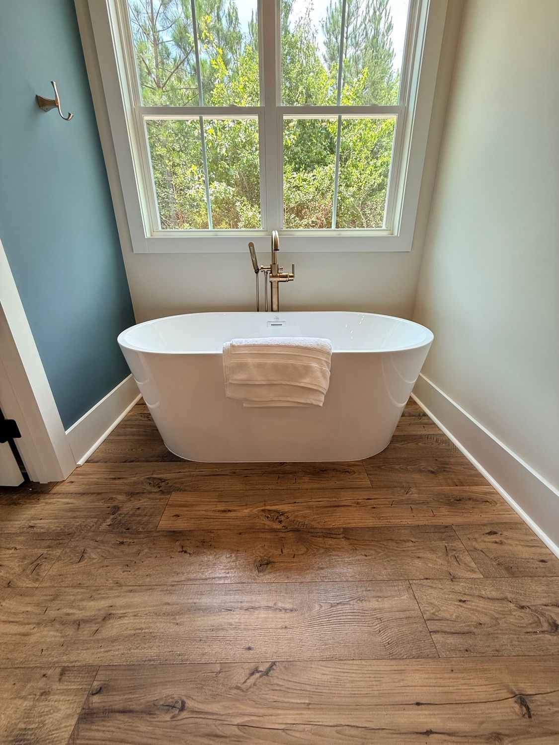 Modern bathroom with freestanding white tub, wood floor, and large window overlooking greenery