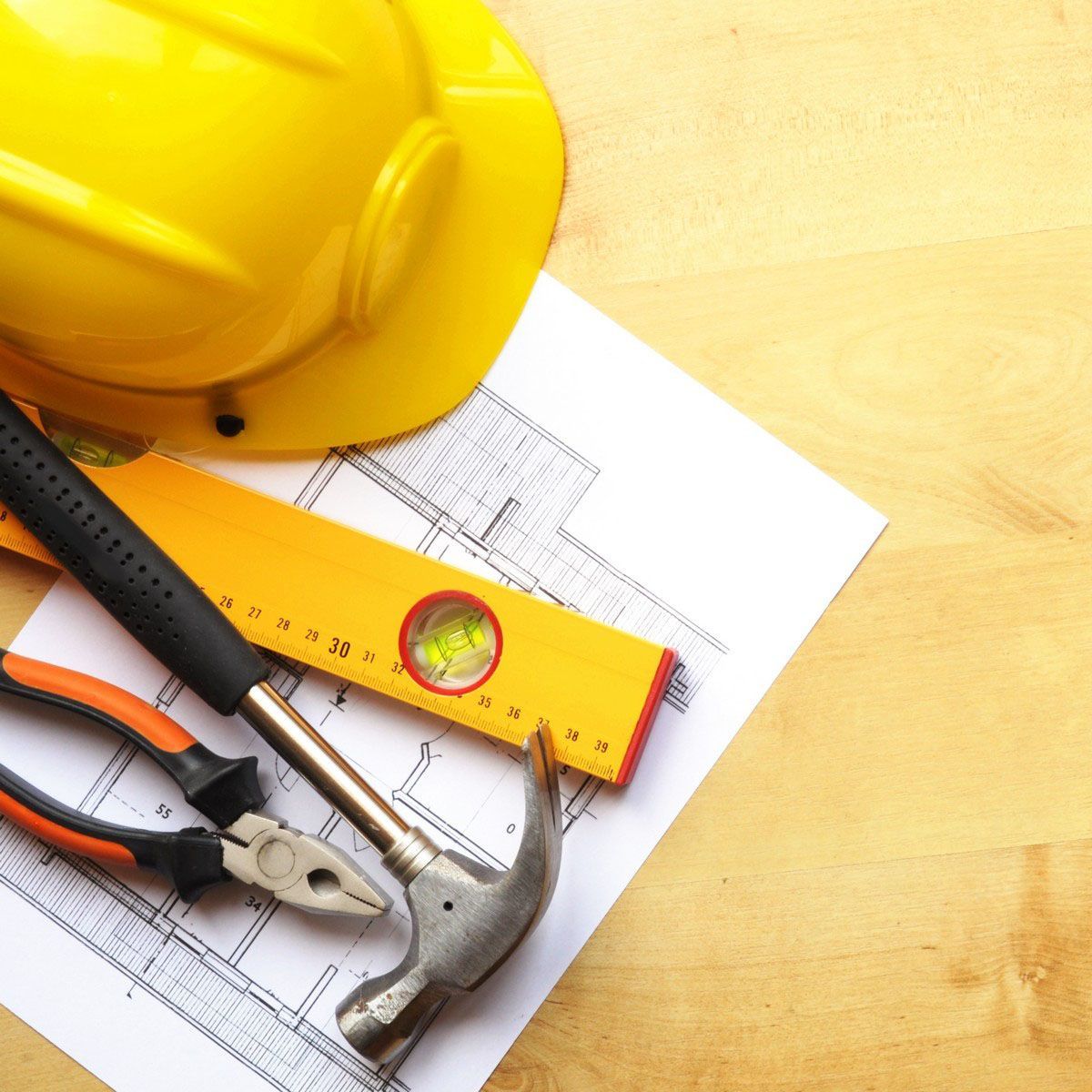 Yellow hard hat, blueprint, hammer, level, and pliers on a wooden surface.
