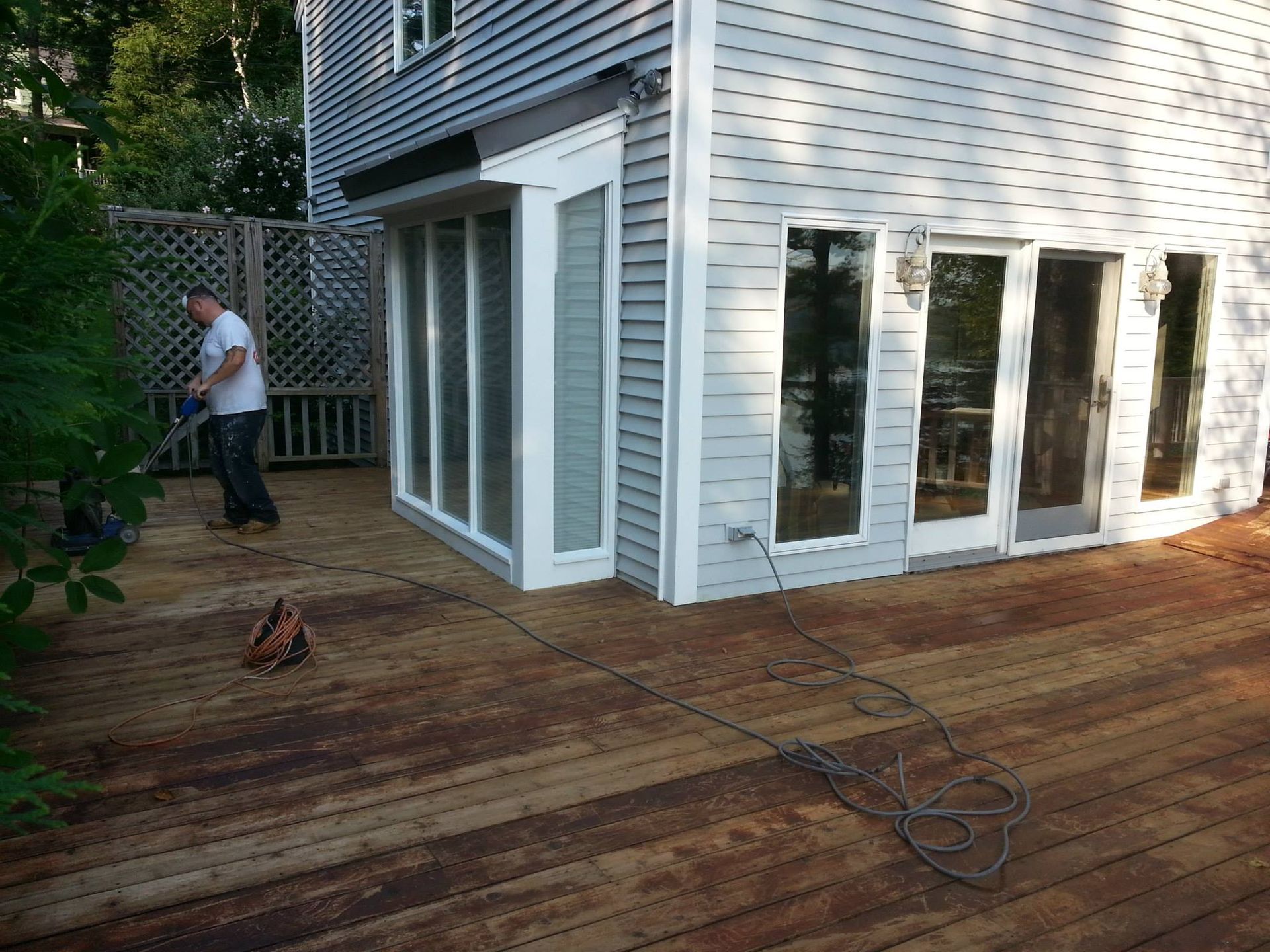 Man using power tool on a weathered wooden deck next to a house with large windows.
