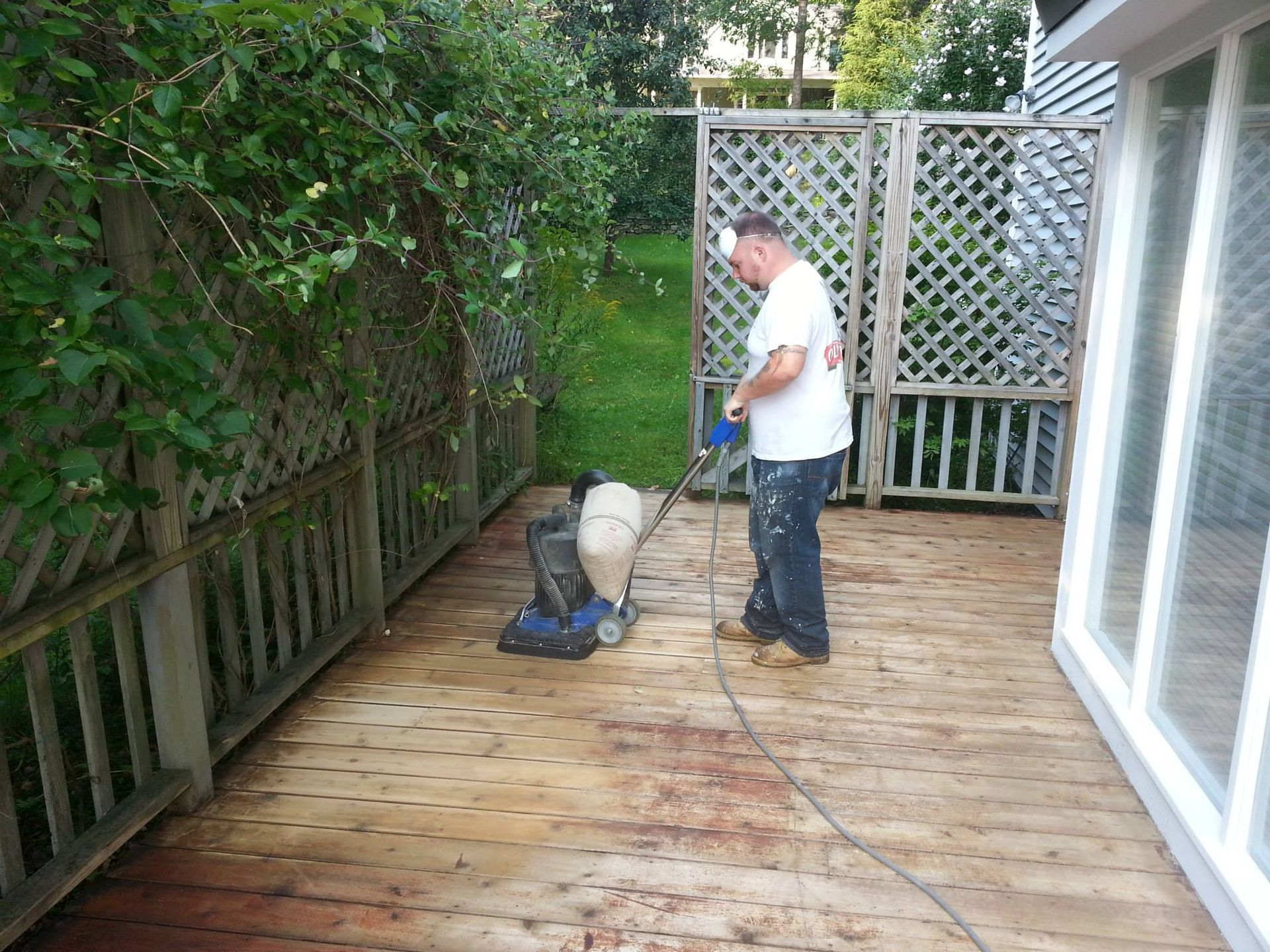 Man power washing a wooden deck with a machine. Green foliage and latticework fence in background.
