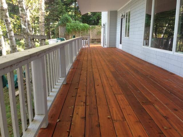 Wooden deck with white railings, against a gray house, trees in background.