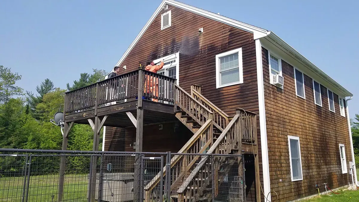 Brown wood house with a deck and stairs, being power washed by a person in the doorway.