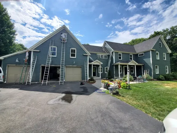 House exterior being painted, blue siding, ladders leaning, two-car garage, driveway, green lawn, clear sky.