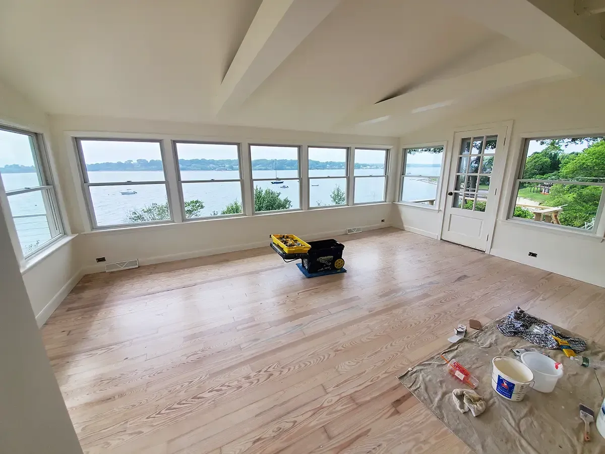 Sunroom with light wood floor and many windows overlooking water. Construction tools and paint supplies present.
