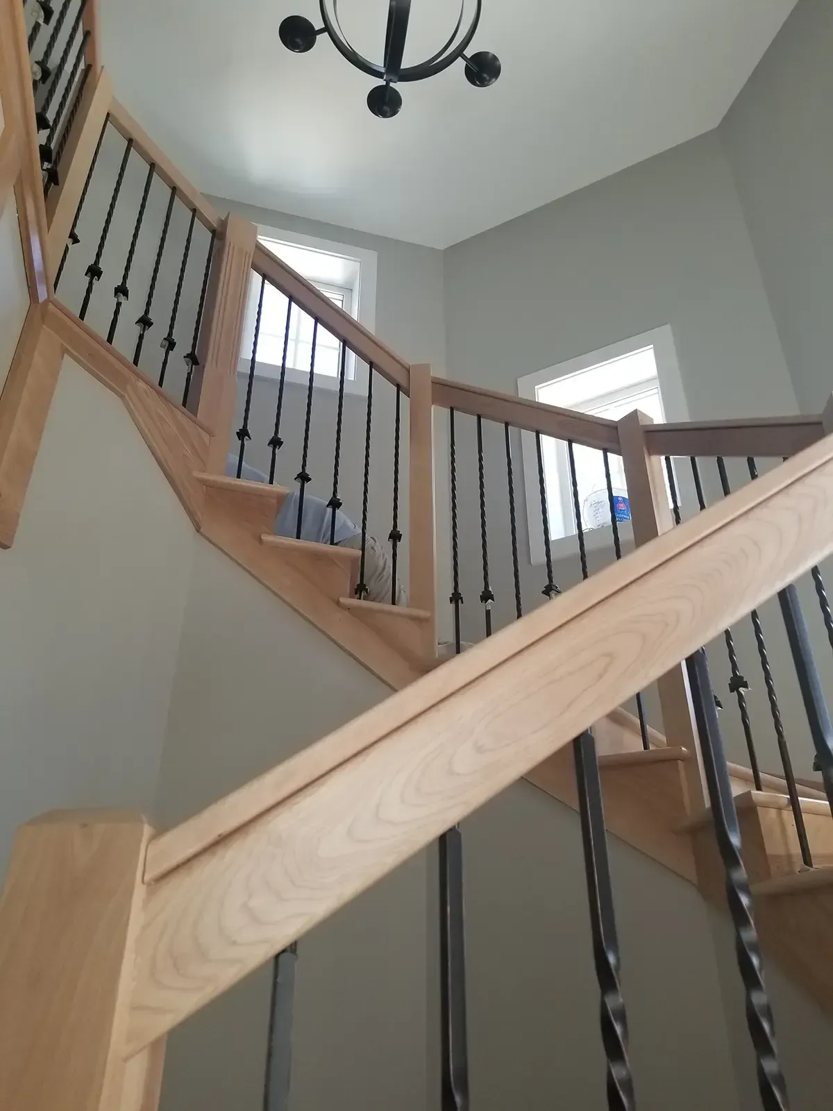 Wooden staircase with black metal balusters, natural wood finish, leading upwards with two windows and a light fixture.
