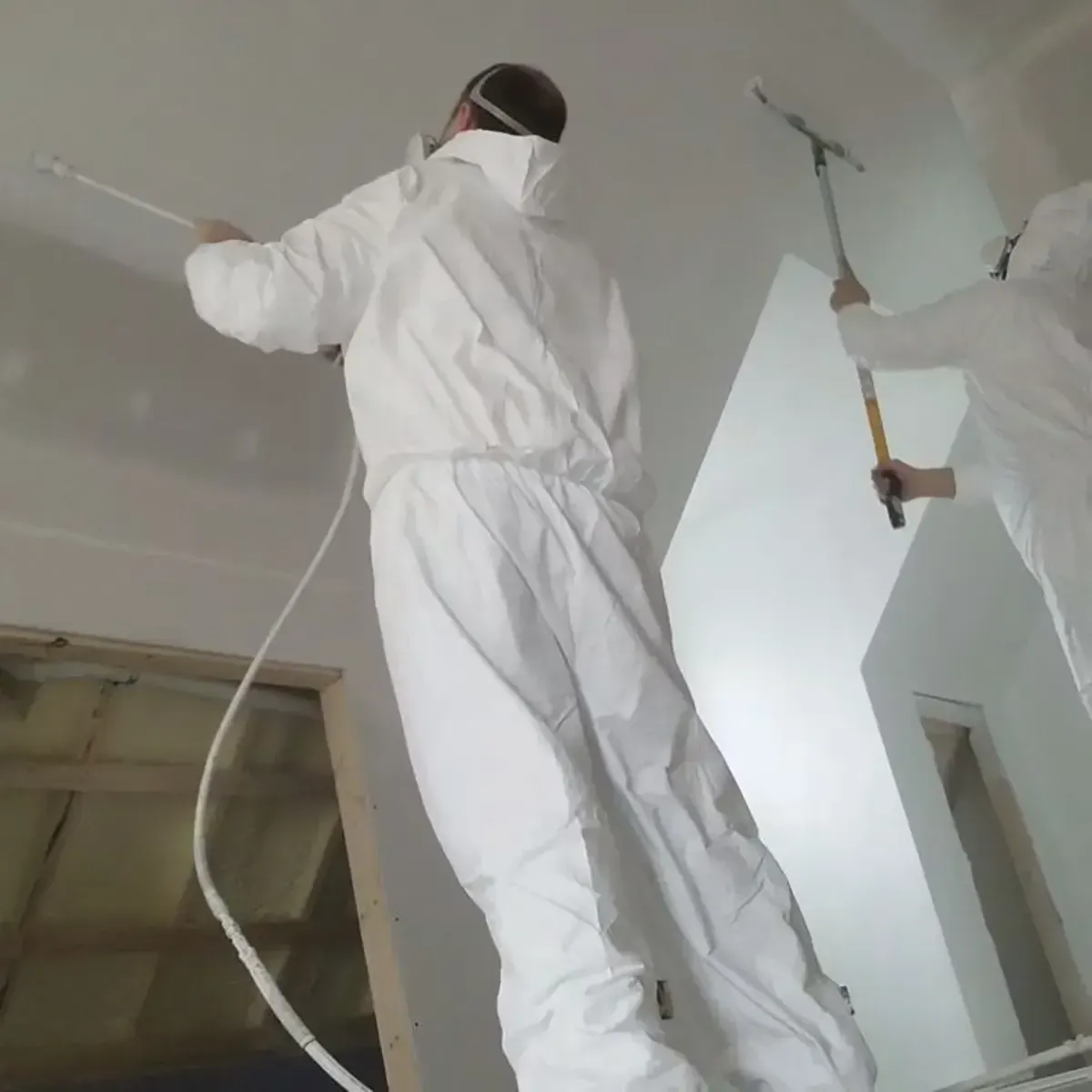 Two people in white protective suits painting a ceiling and wall with spray equipment and a roller in an interior space.