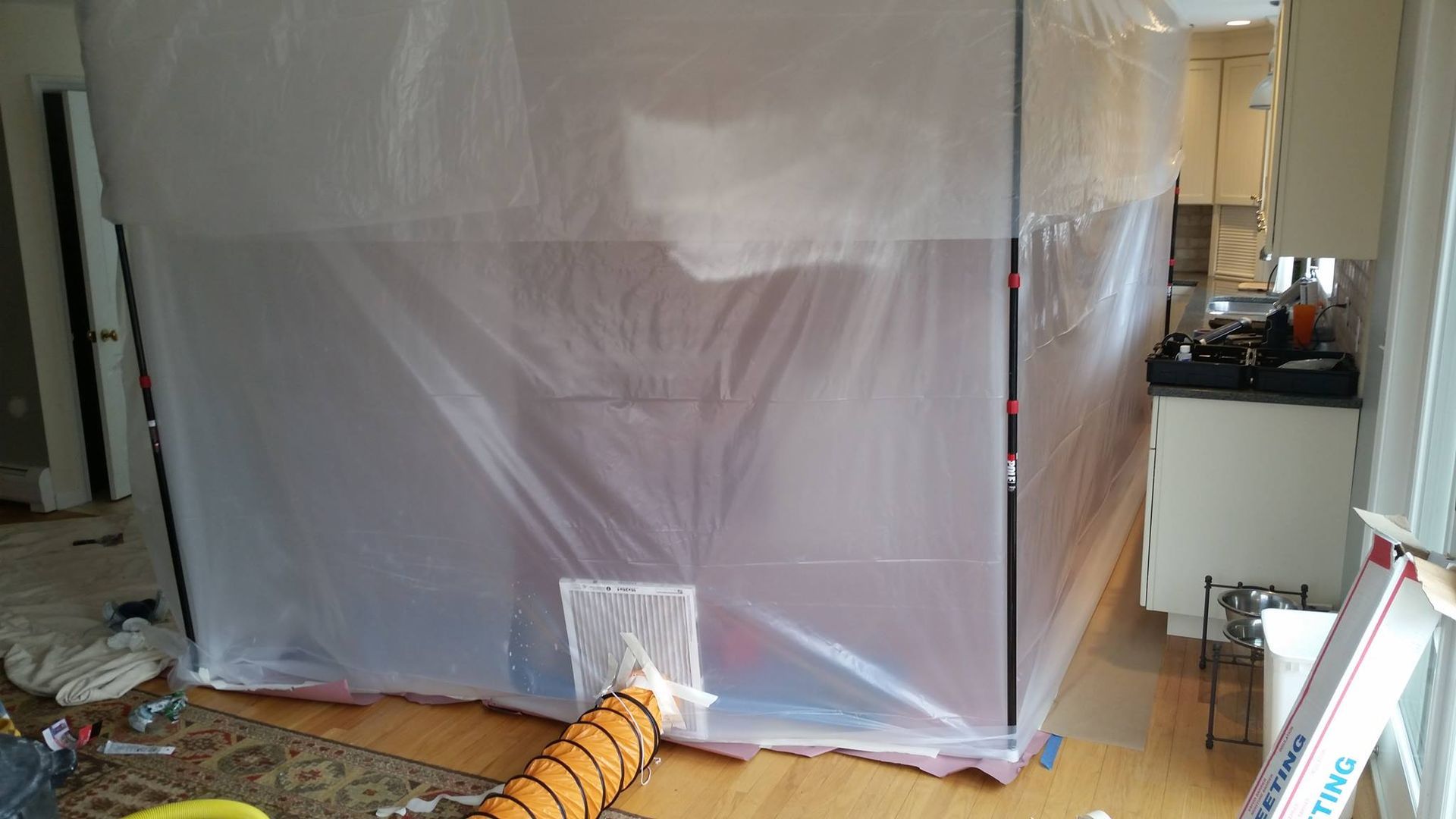 A room is encased in plastic sheeting. An orange hose extends from a rectangular opening near the bottom.