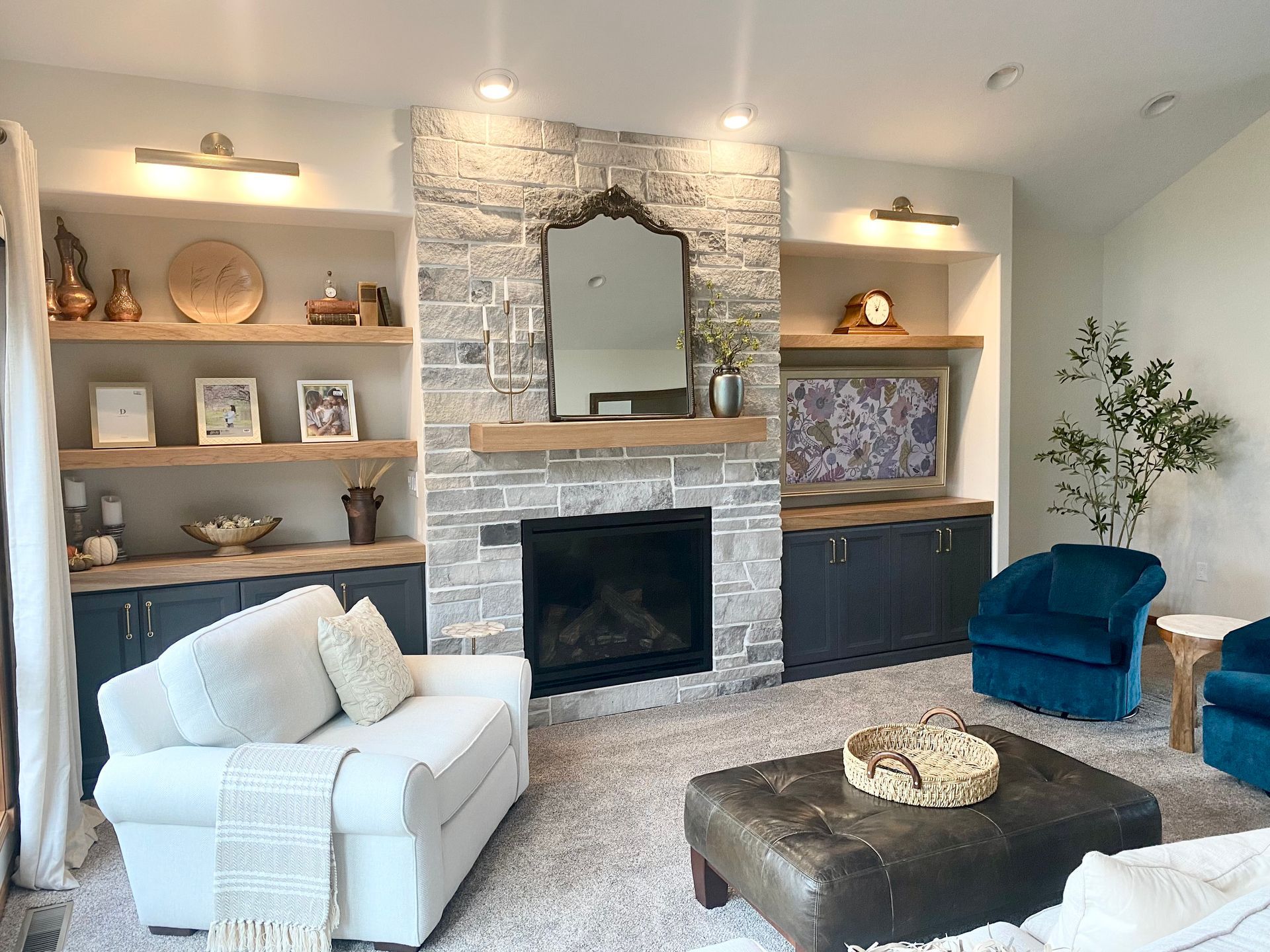 A cozy living room with a stone fireplace, built-in shelving, a white armchair, and deep blue velvet chairs.