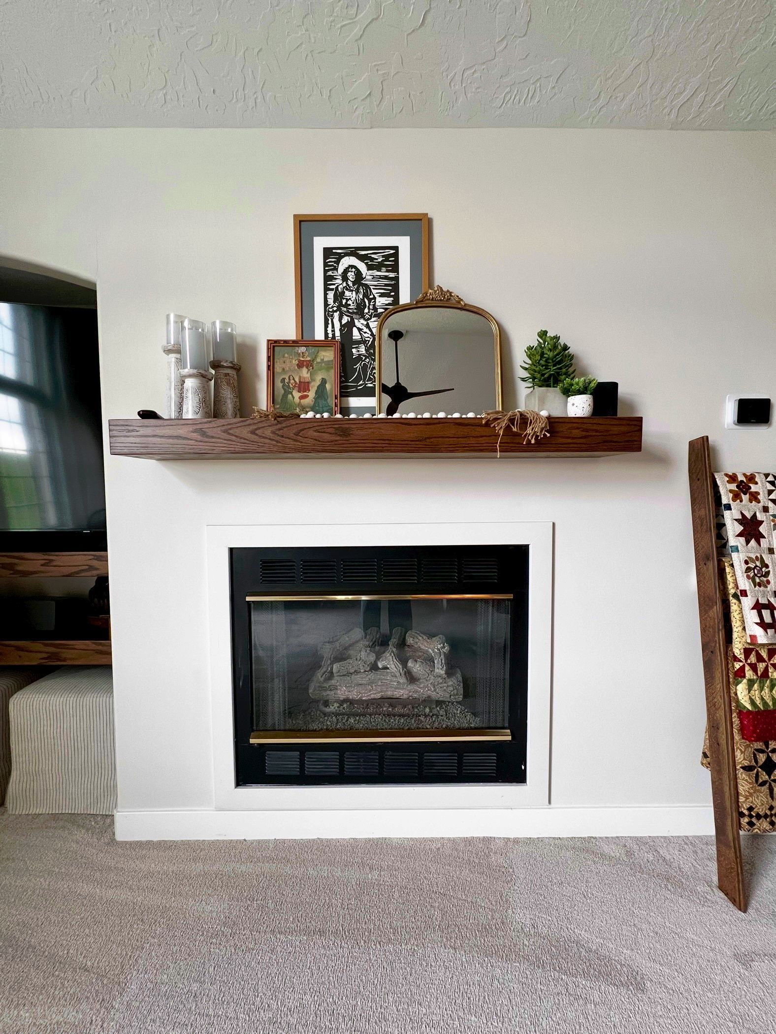 A living room fireplace with a dark wood mantel, decorated with framed art, a small gold mirror, and greenery.