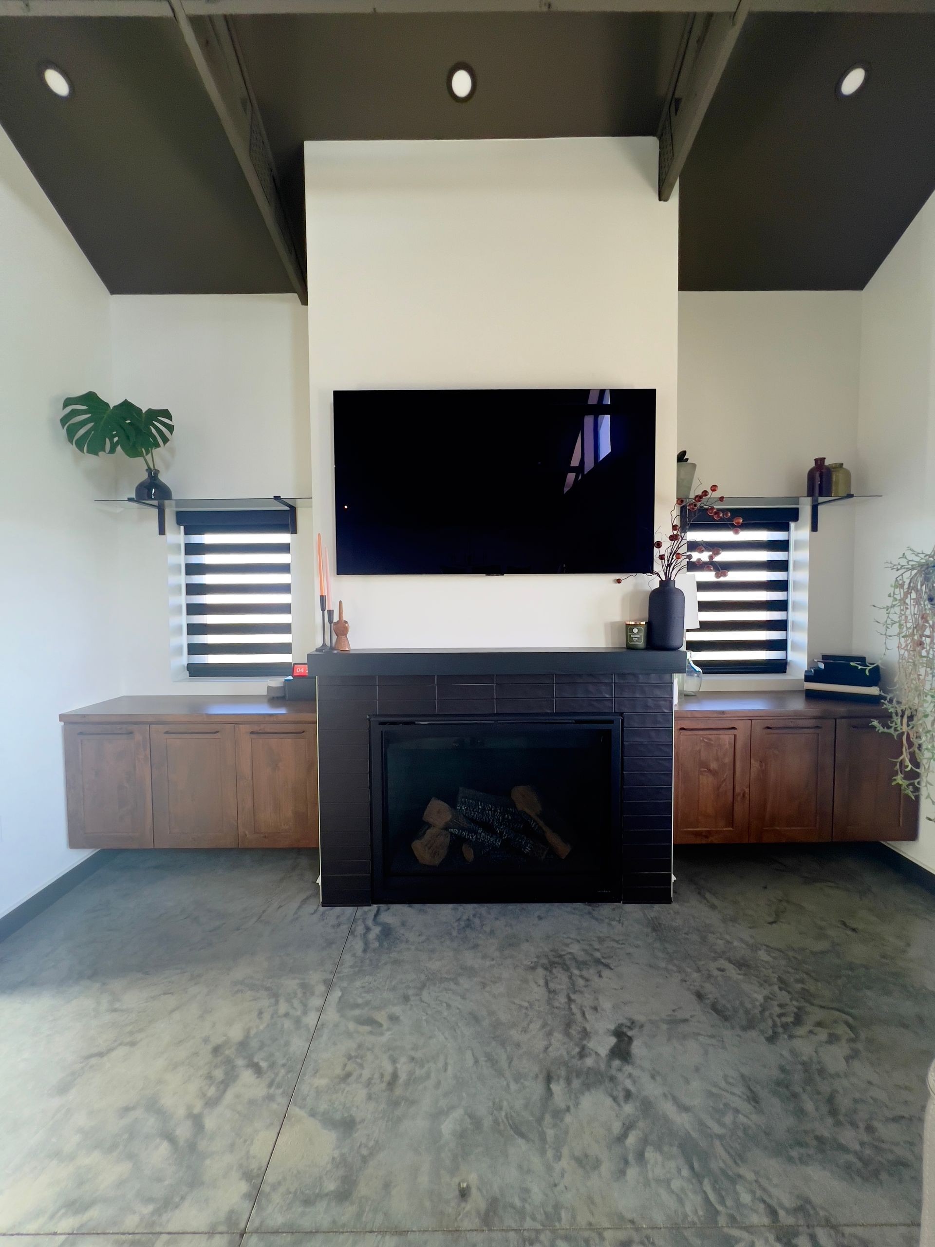A fireplace with a mounted TV above, flanked by wooden cabinets and windows with black blinds in a room with dark ceilings.