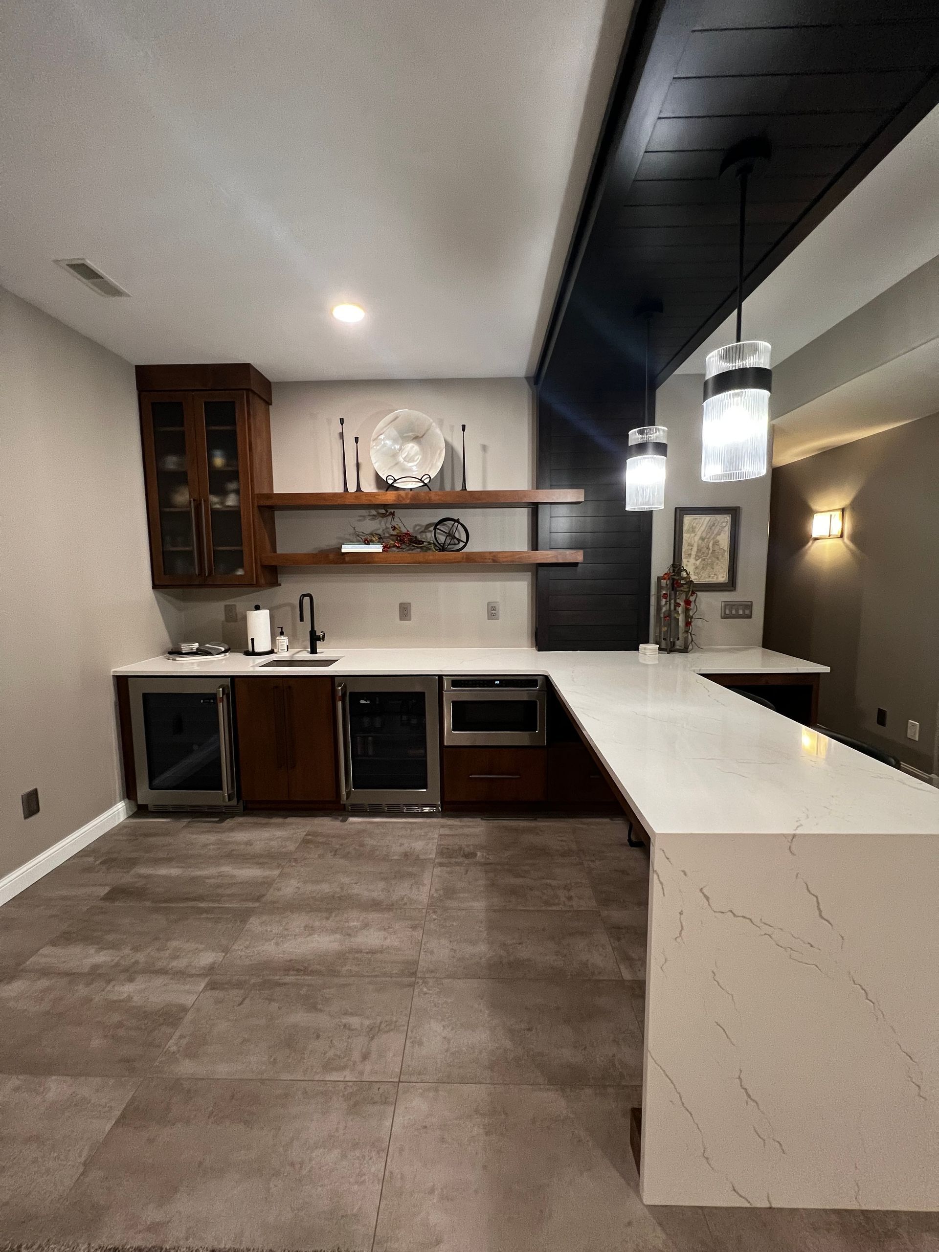 Modern wet bar with dark wood cabinets, white quartz counters, open wood shelving, and two pendant lights above the bar.